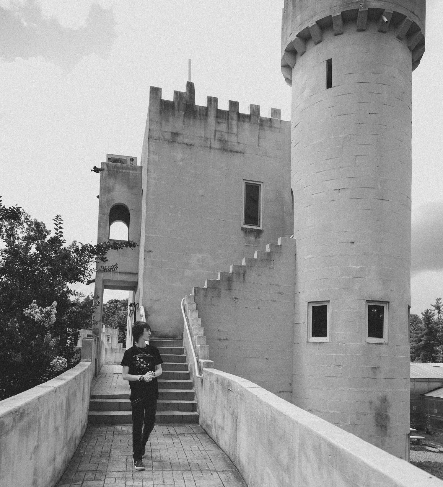 Black and white photo of a person walking along a stone walkway toward a castle-style building with a round tower and crenellated walls.