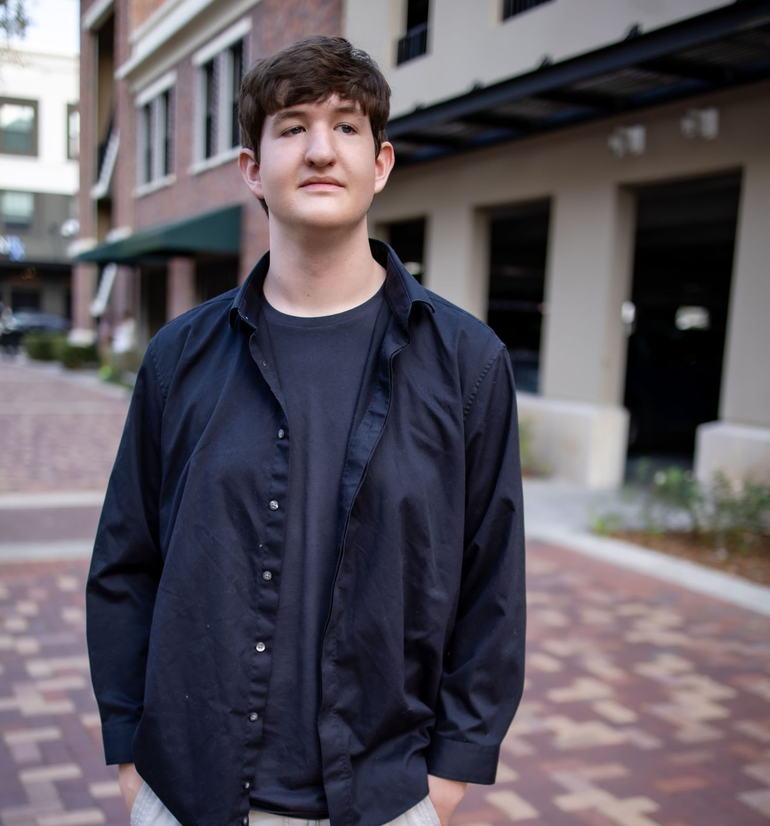 A person wearing a black button-up shirt poses in front of a brick building in a casual outdoor setting.