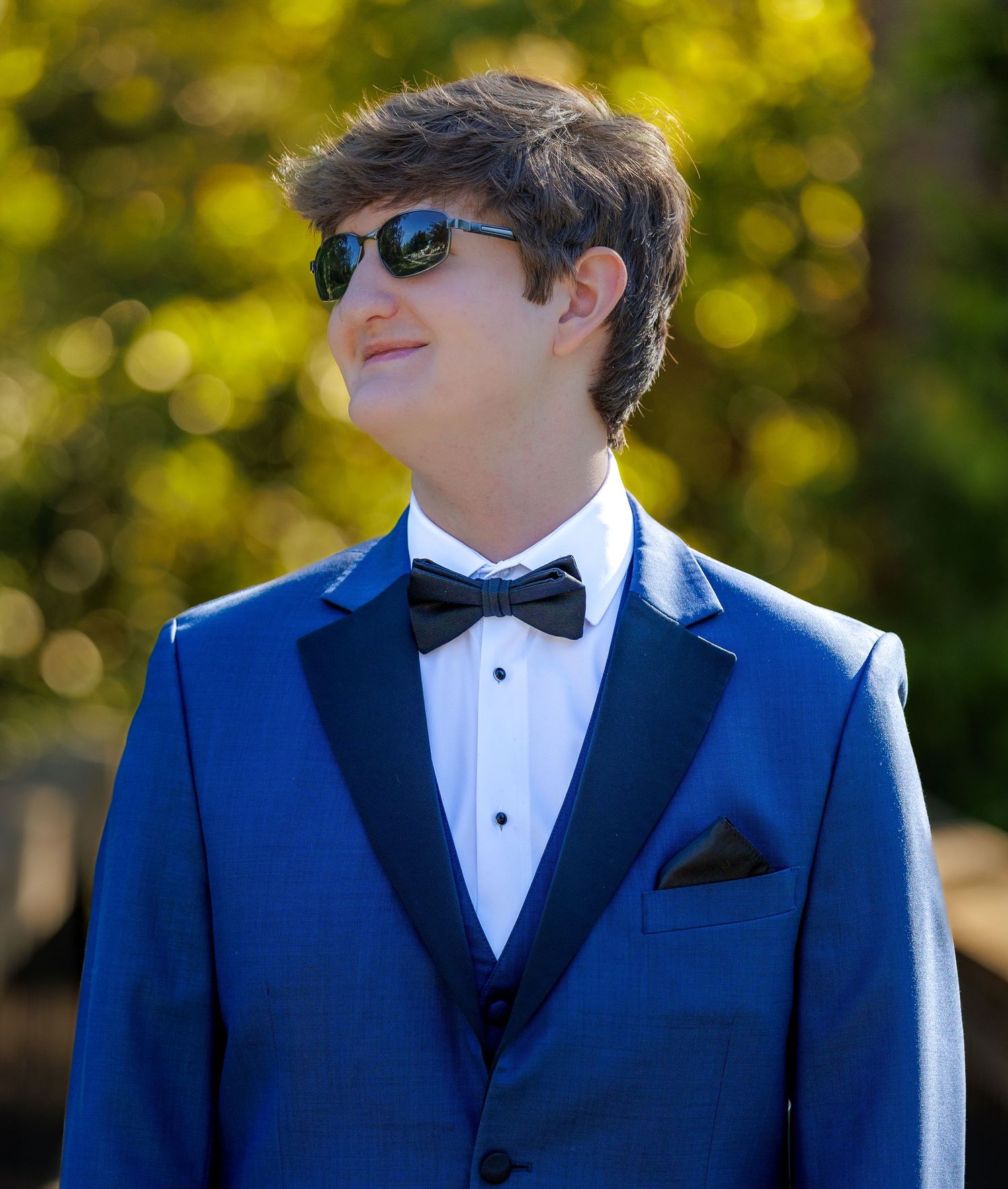 Young man in blue tuxedo with black bow tie and sunglasses posing outdoors for formal event photo.