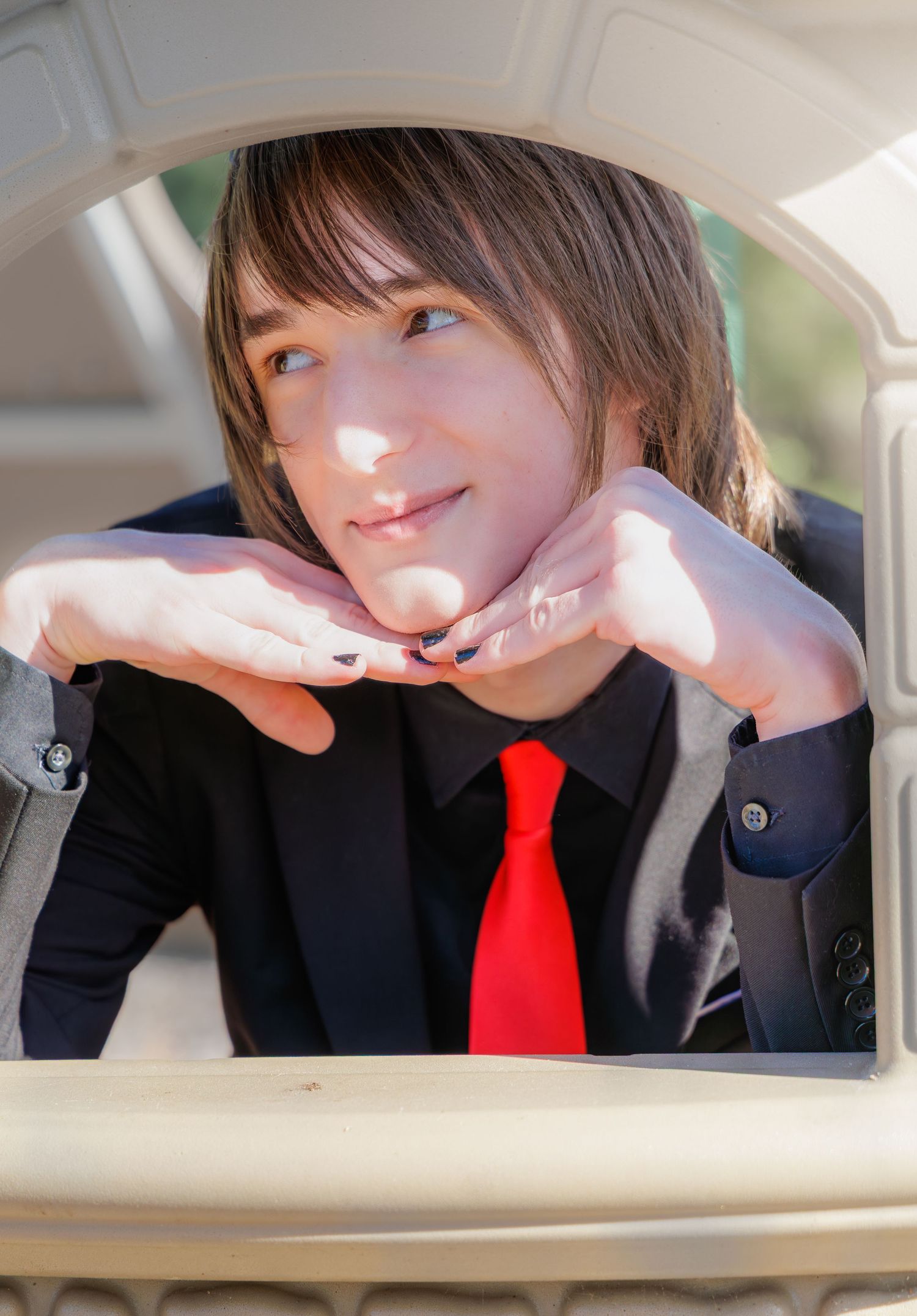 Young person in black suit and red tie resting chin on hands, smiling outdoors in natural light.