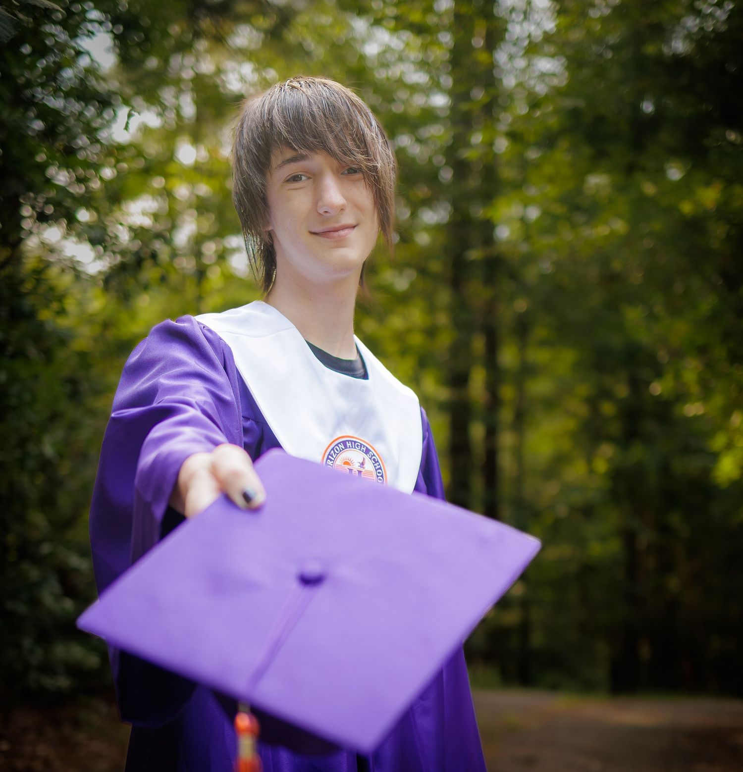 Smiling graduate in a purple gown and white stole extends a purple mortarboard toward the camera in a sunlit forest.