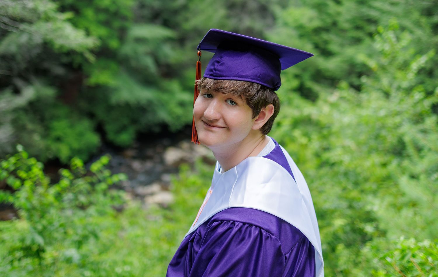 Graduate in a purple cap, gown, and white stole smiles over their shoulder while posing among lush green foliage outdoors.