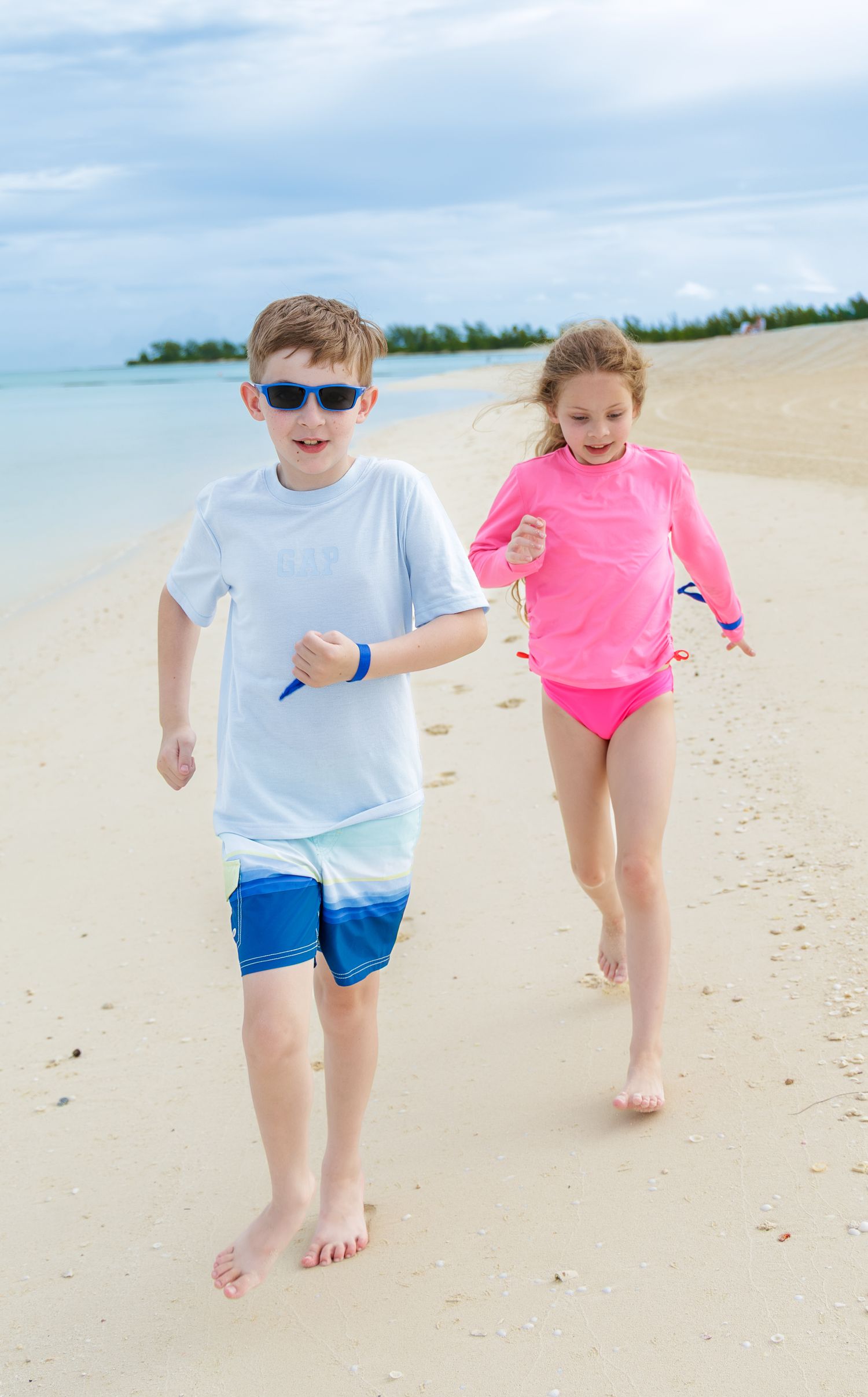 Two smiling children in swimwear, a boy in sunglasses and a girl in pink, run barefoot along a sandy tropical beach by calm turquoise water.
