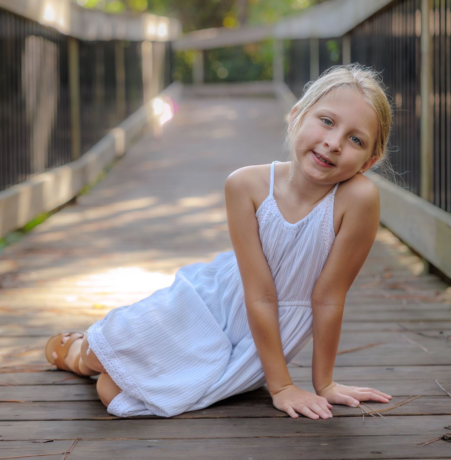 Young girl with blonde hair in a white sundress sits smiling on a sunlit wooden boardwalk surrounded by green trees.