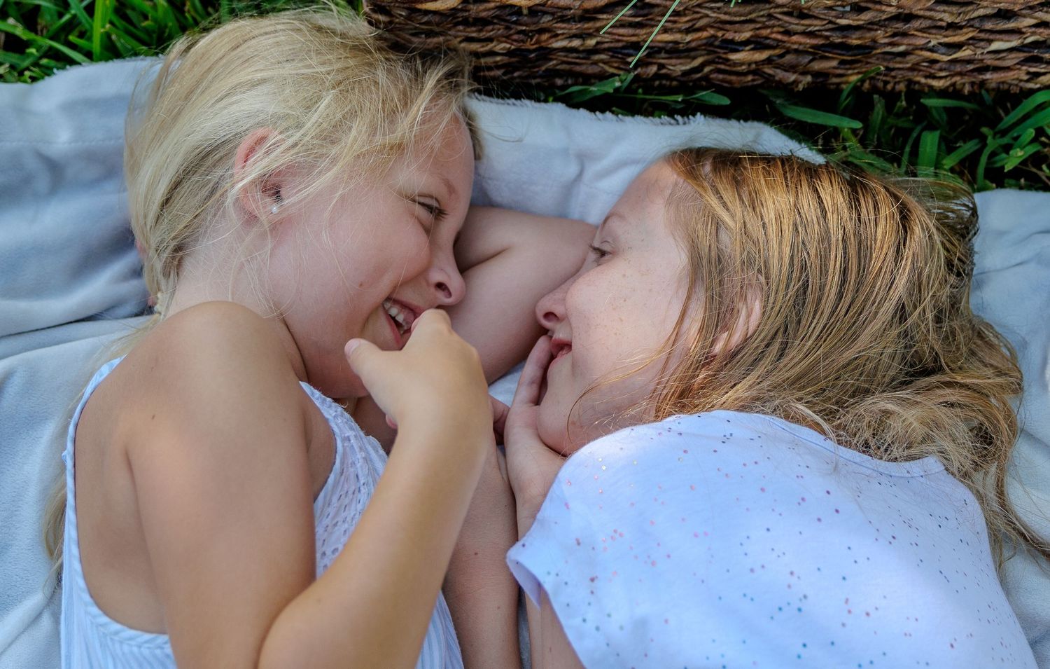 Two young blonde girls lie side by side on a blanket outdoors, laughing and smiling together near a wicker basket on the grass.