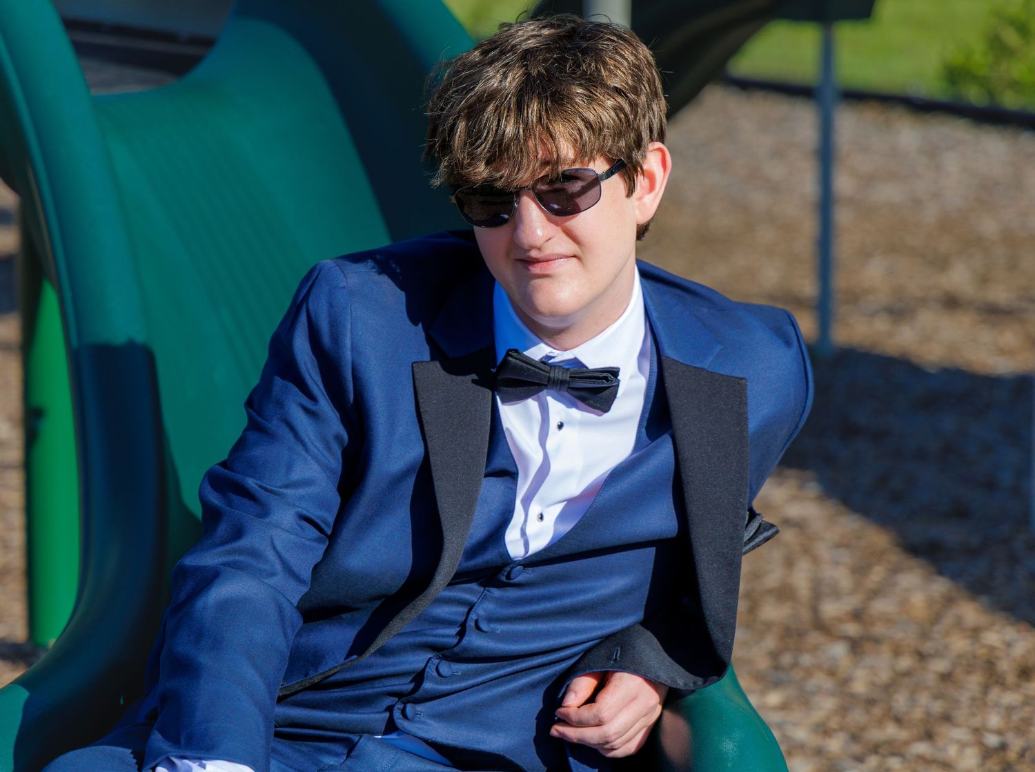 Young boy in blue tuxedo with bow tie and sunglasses sitting on playground slide.