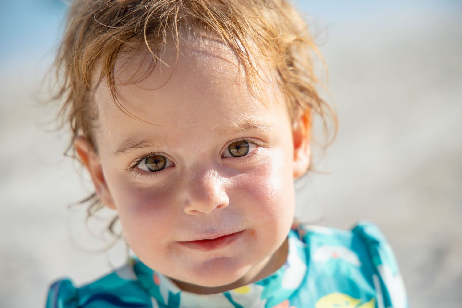 A small child with curly red hair and bright eyes smiles while wearing a turquoise shirt.