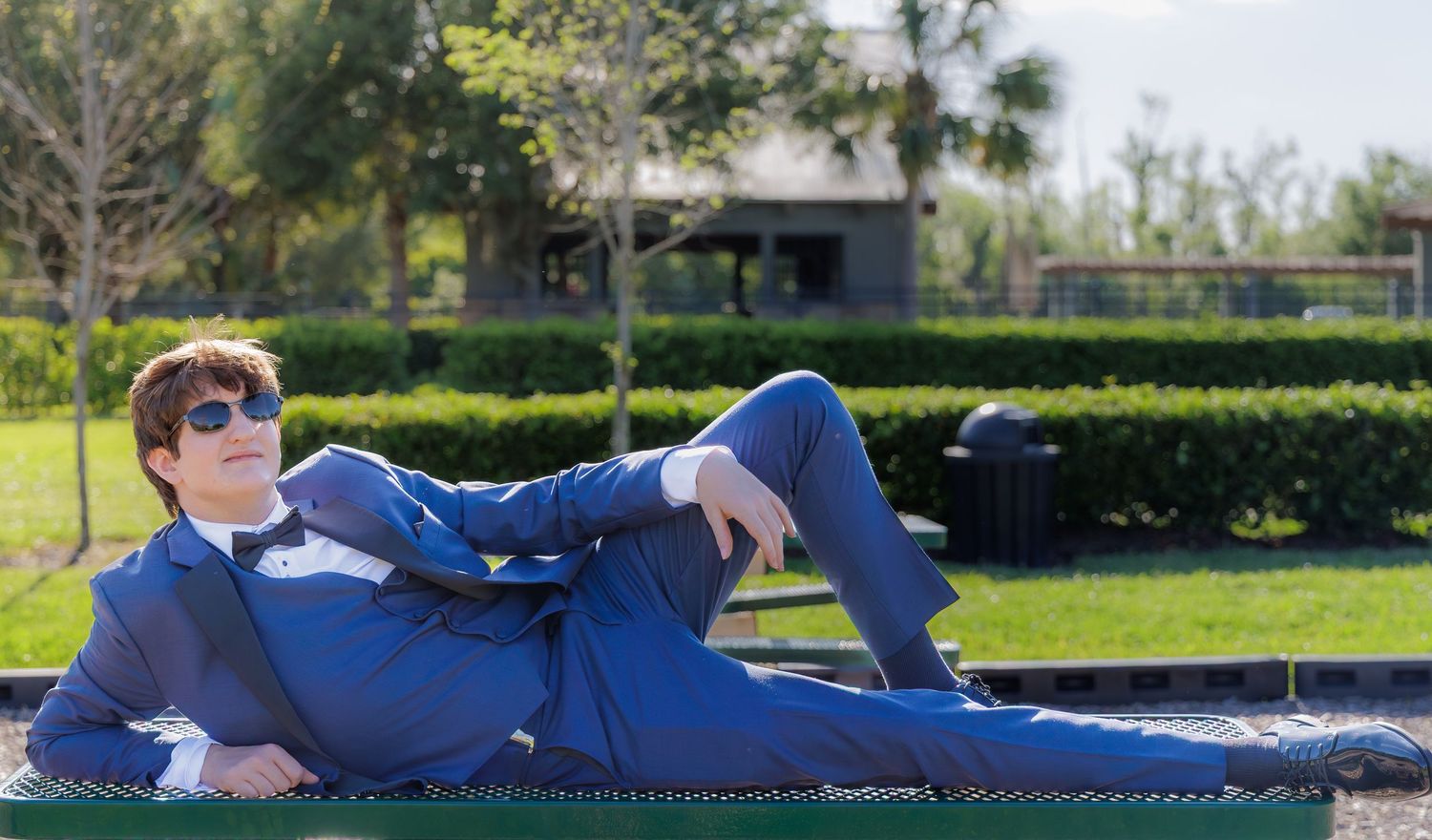 Young man in blue suit and sunglasses relaxing on a bench in a lush green park on a sunny day.