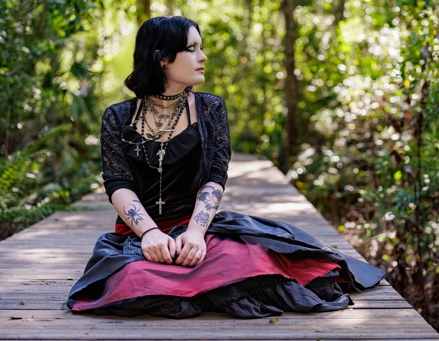 Woman in gothic attire with layered cross necklaces and tattoos sits on a wooden boardwalk in a sunlit forest wearing a red and black skirt.
