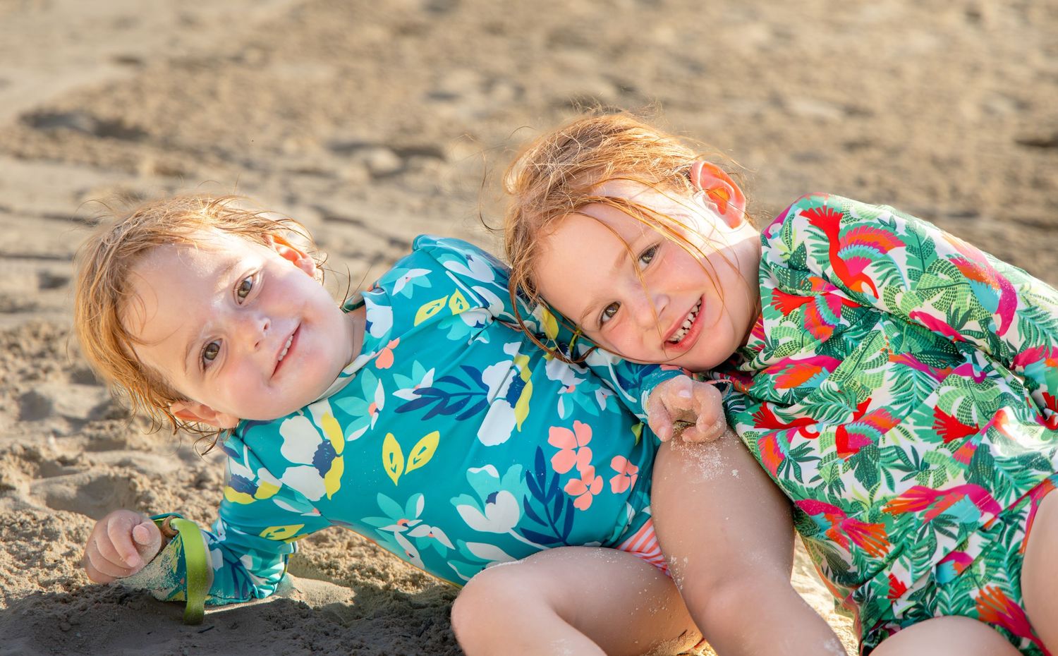 Children in colorful floral dresses play together on a sandy beach during a sunny day.