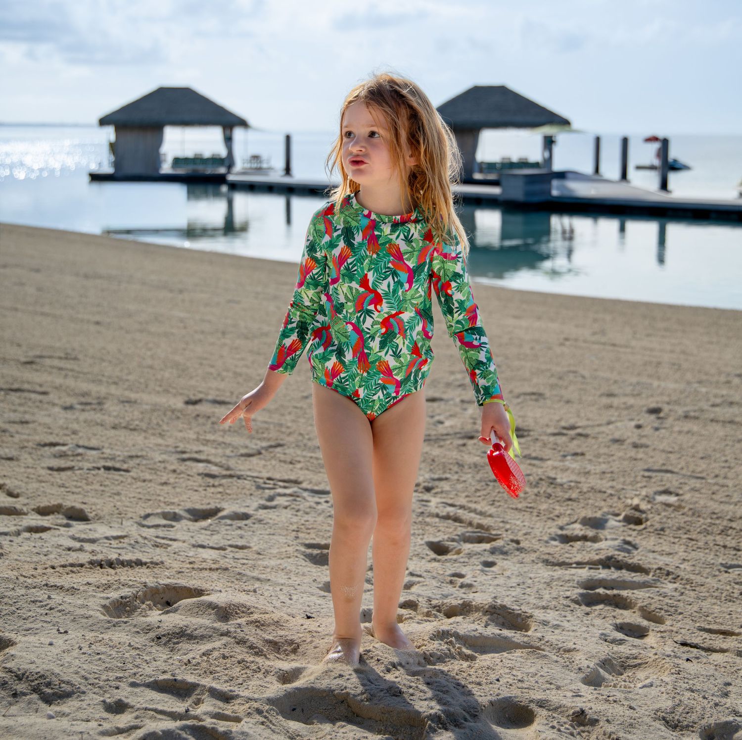 Young girl in a green long-sleeve swimsuit with red parrot print stands barefoot on a sandy beach with overwater huts behind her.