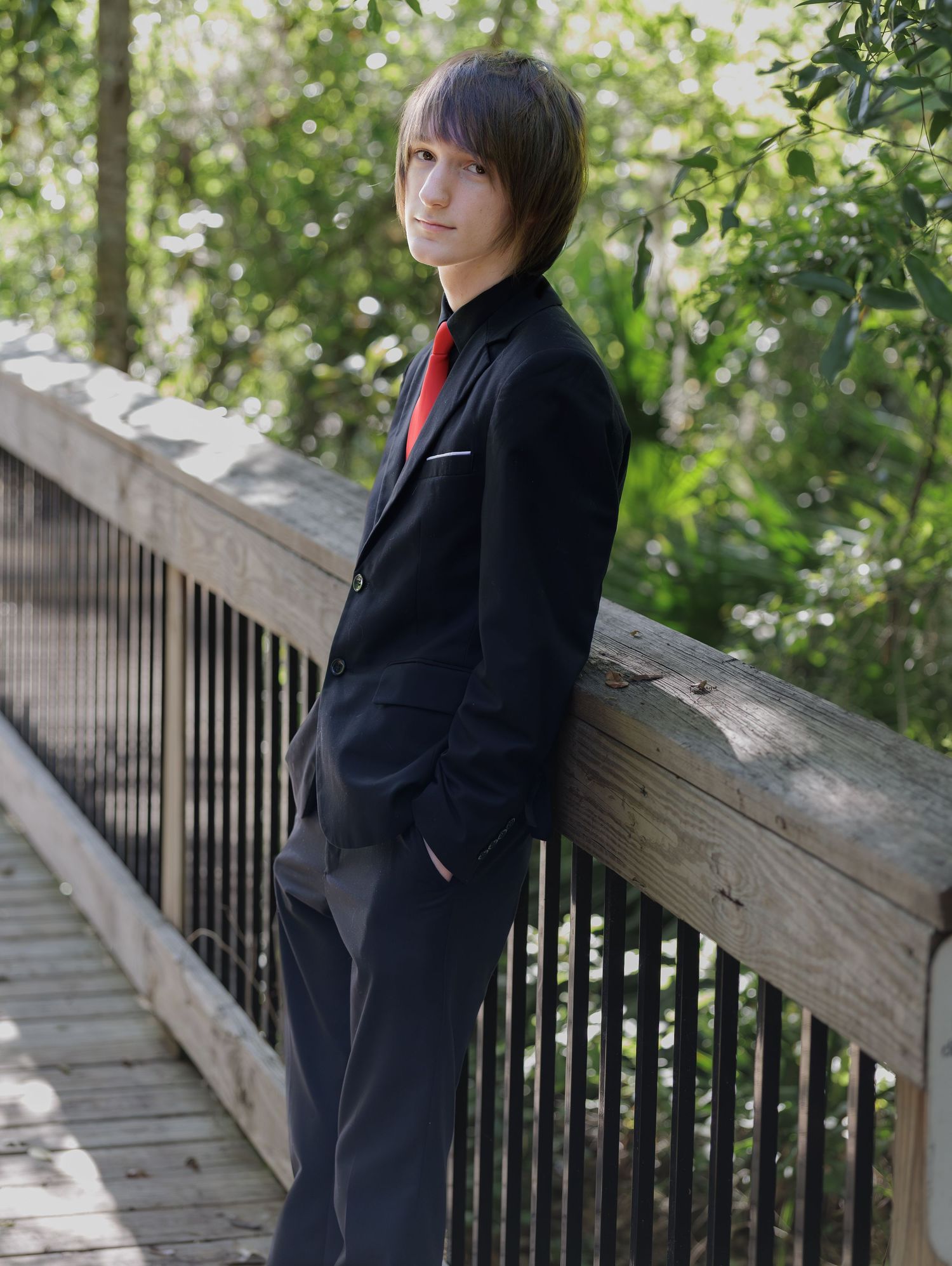 A young man in a black suit with red tie leans against a wooden railing on a bridge surrounded by greenery.