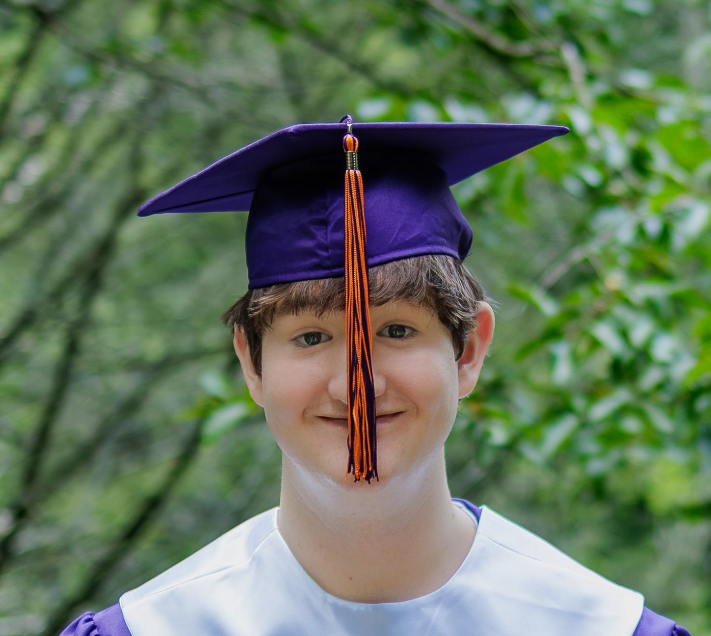 Smiling graduate in a purple cap with an orange tassel and white gown posing outdoors in front of green leafy trees.