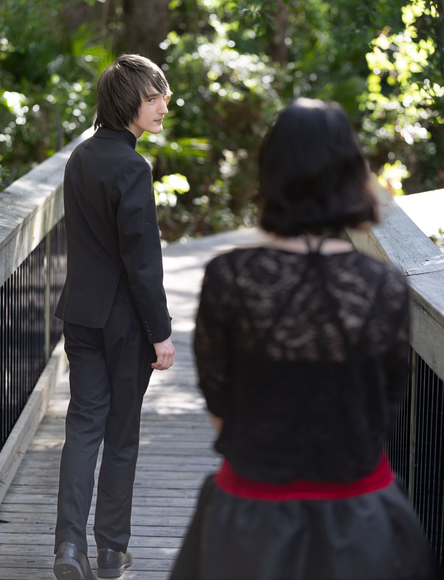 A young man in a black suit stands on a wooden bridge, looking back at a woman in gothic black lace dress.