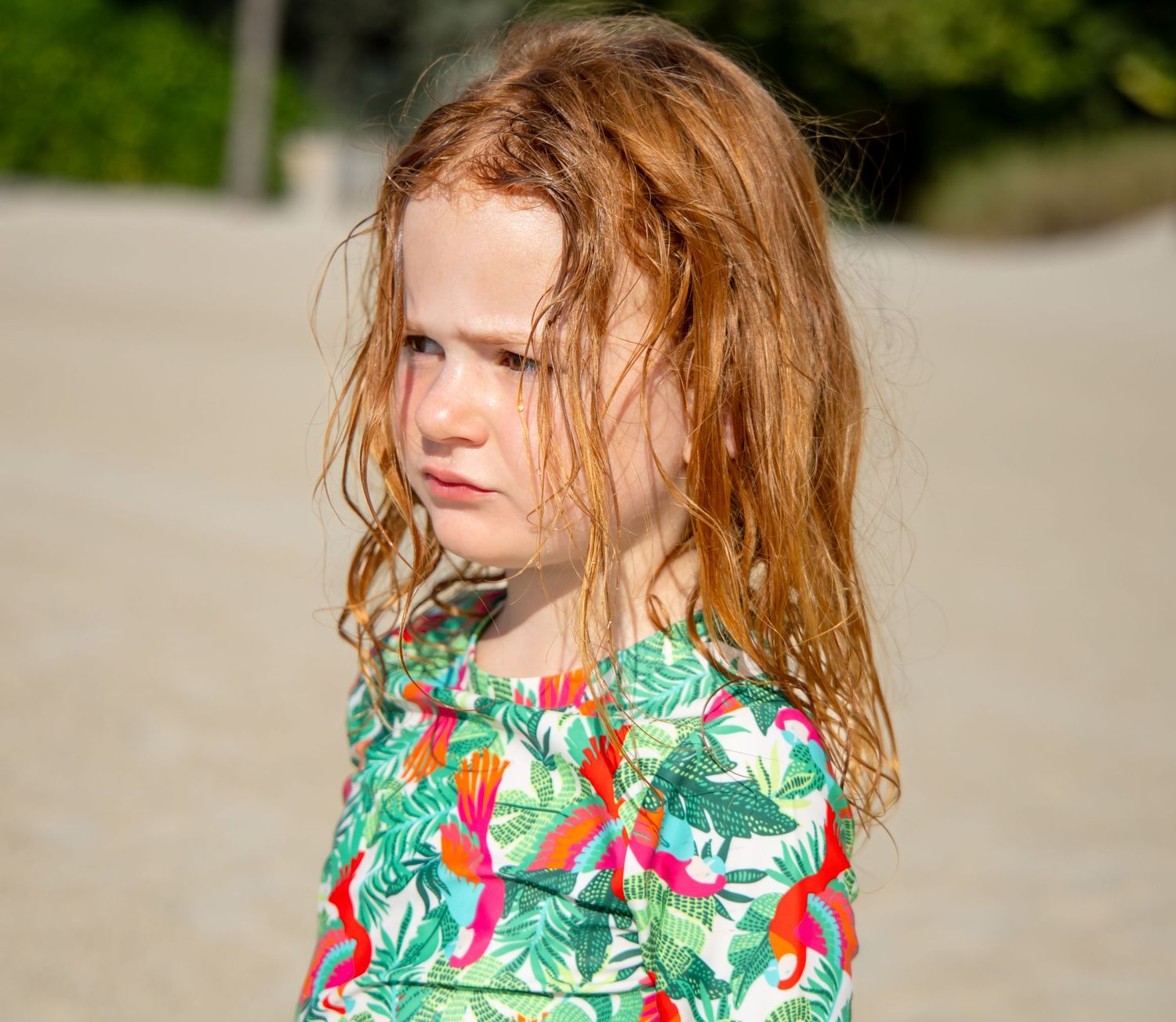 Young child with wet ginger hair wearing a colorful tropical print swimsuit stands on a beach.