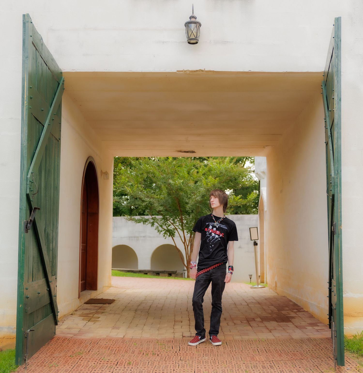 Young man in a dark graphic t-shirt and black jeans stands in a whitewashed archway with open green wooden doors, looking upward.