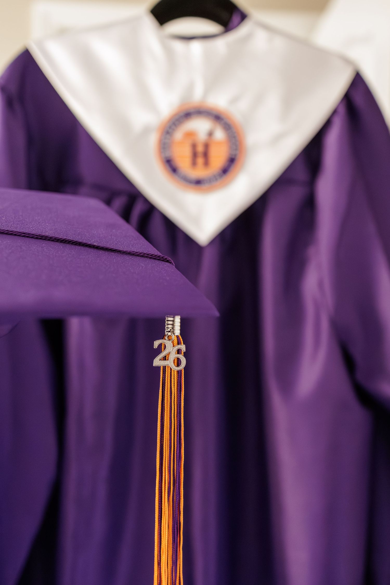 Purple graduation gown with white hood featuring an H emblem and a mortarboard cap with gold tassel charm reading 28.