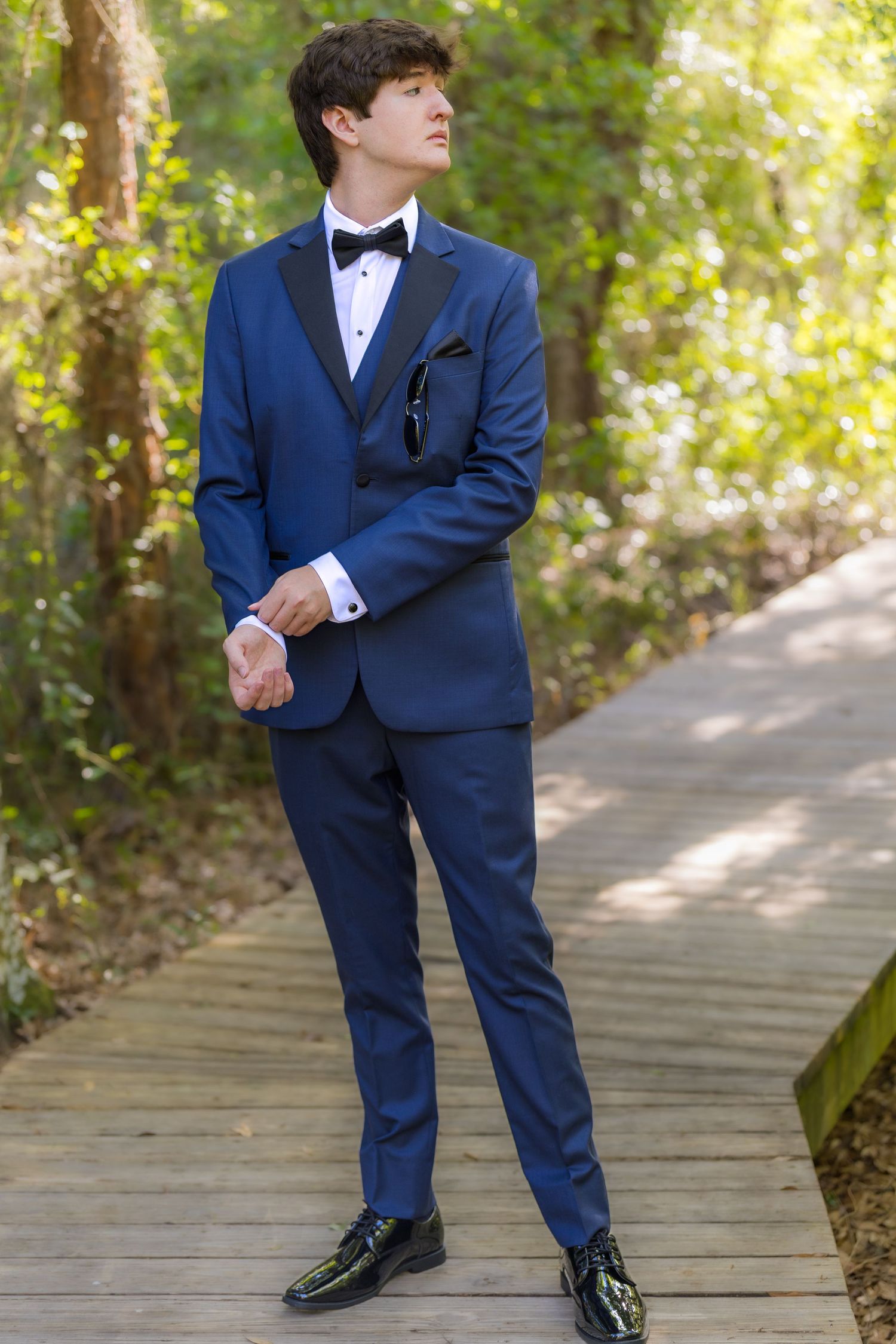 Young man in navy blue tuxedo with black lapels and bow tie standing on wooden path surrounded by lush greenery.