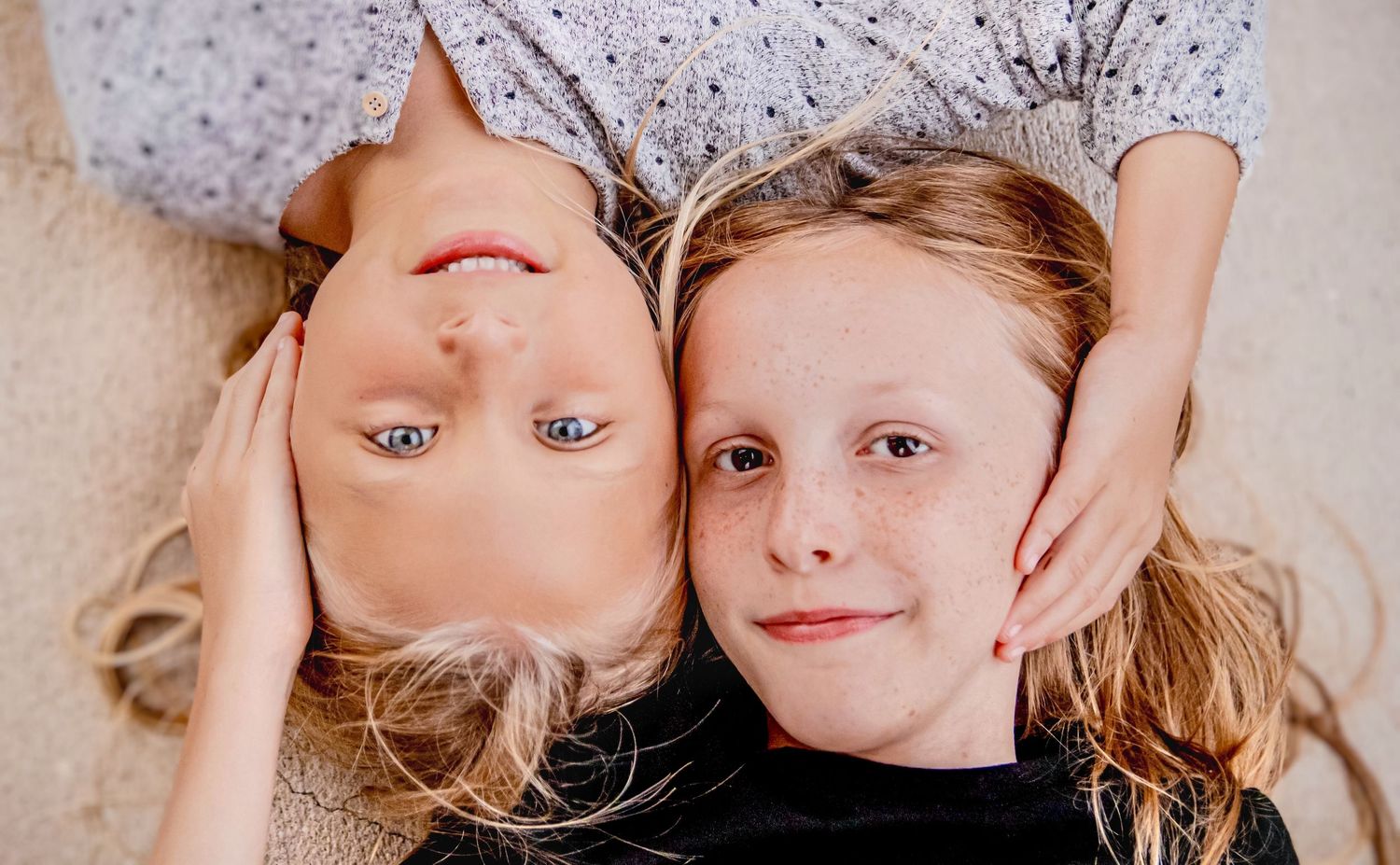 Three playful children lying on floor looking up at camera with silly expressions and blonde hair.