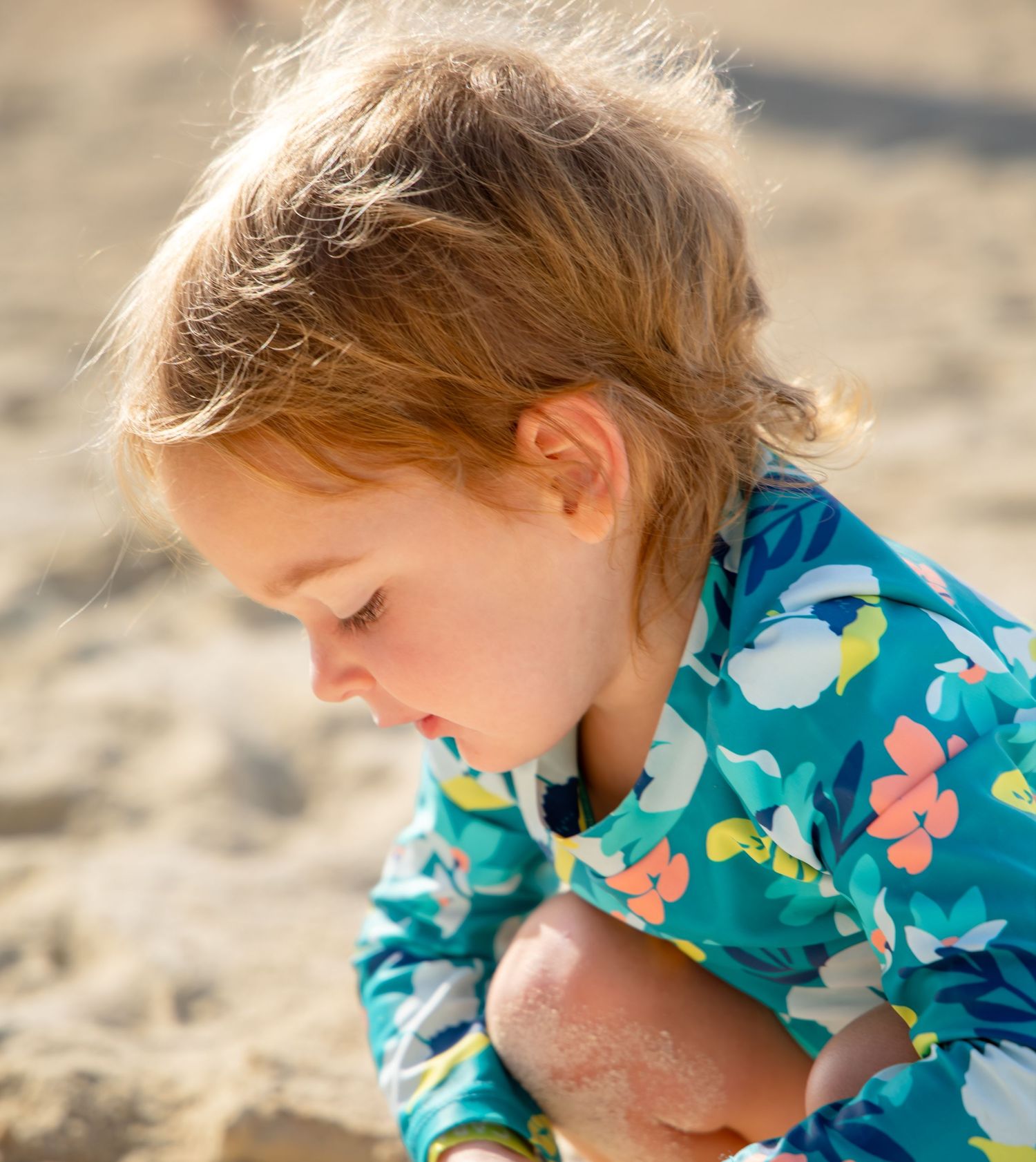Child in turquoise floral swimwear plays in sand at beach on sunny day.