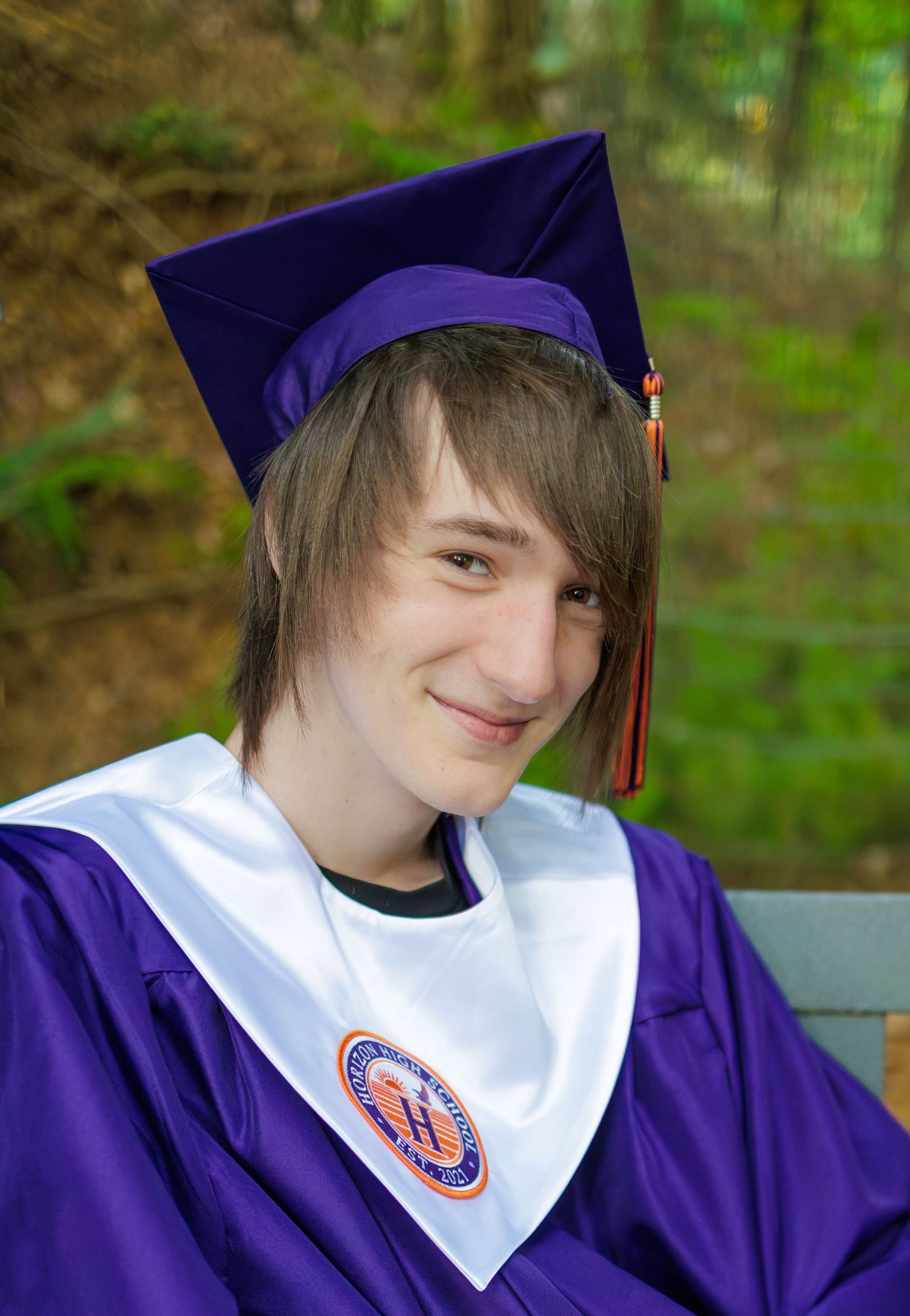 Smiling graduate in a purple cap and gown with a white stole and orange tassel sits outdoors on a bench in front of green trees.