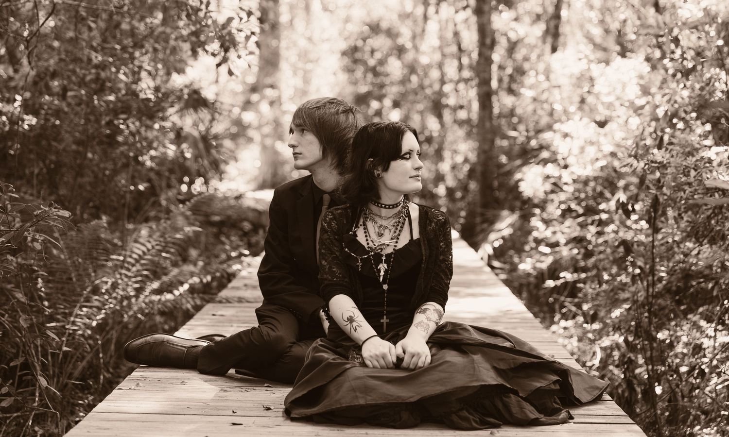Black and white photo of two people sitting closely on a wooden bridge surrounded by lush forest greenery.