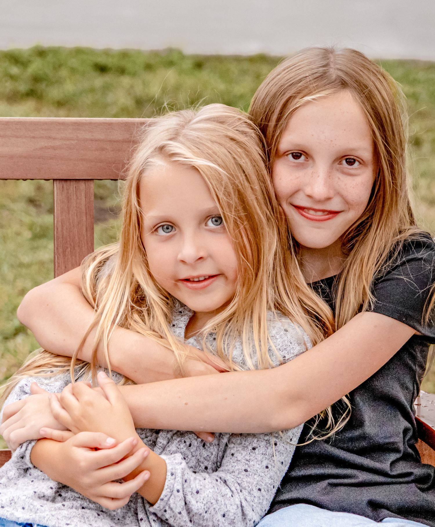 Two young children with long blonde hair embrace while sitting together outdoors.