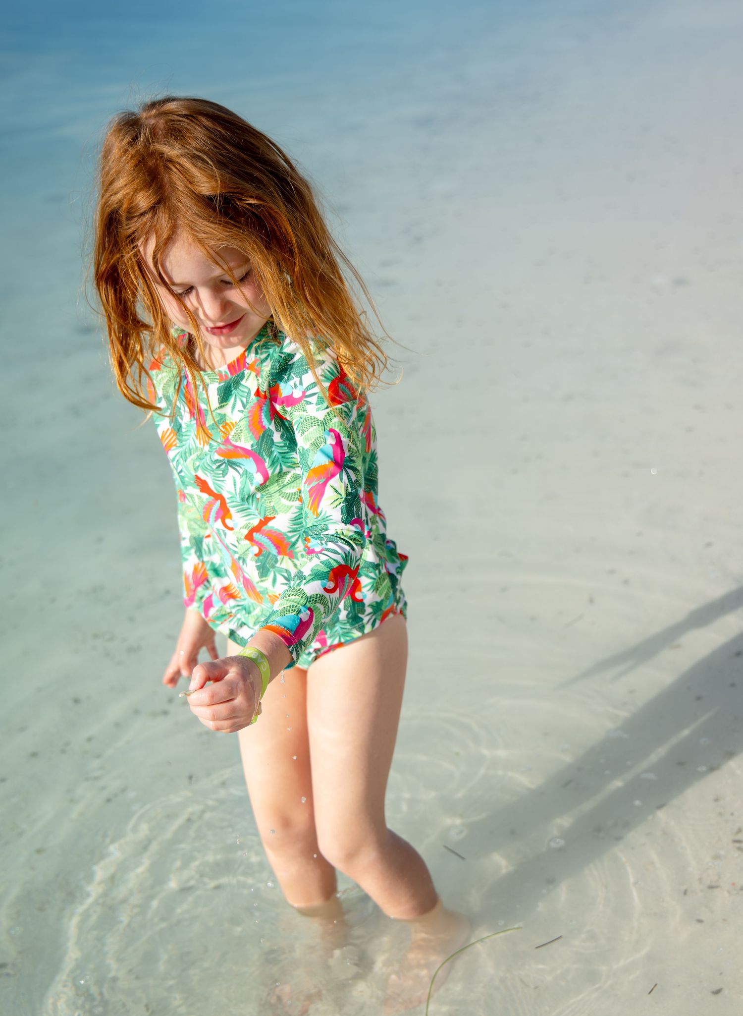 A person in a colorful floral swimsuit plays in shallow turquoise ocean water on a sunny beach.