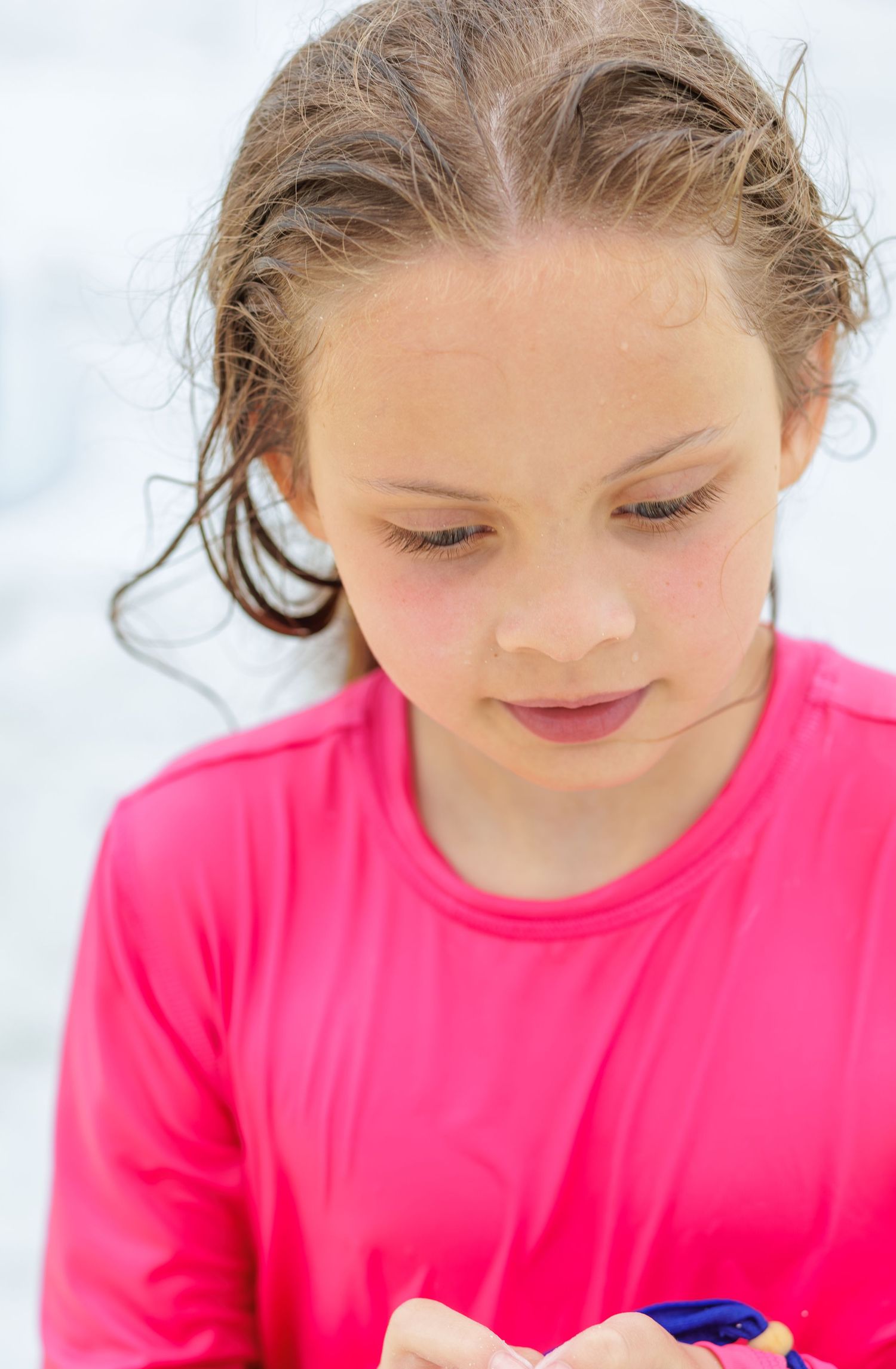 Young girl with wet hair in a bright pink long-sleeve shirt looks down at her hands against a soft white background.