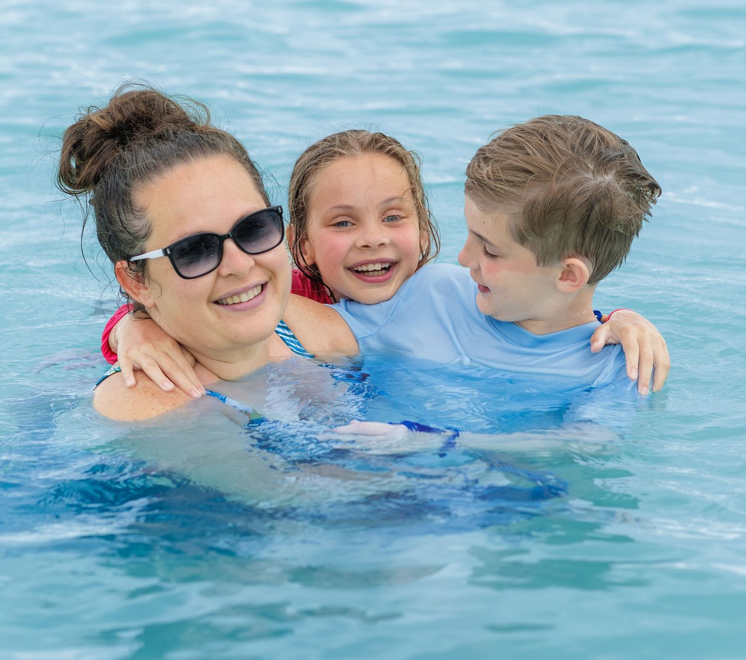 Smiling woman in sunglasses hugs two happy children while playing together in a bright blue swimming pool.