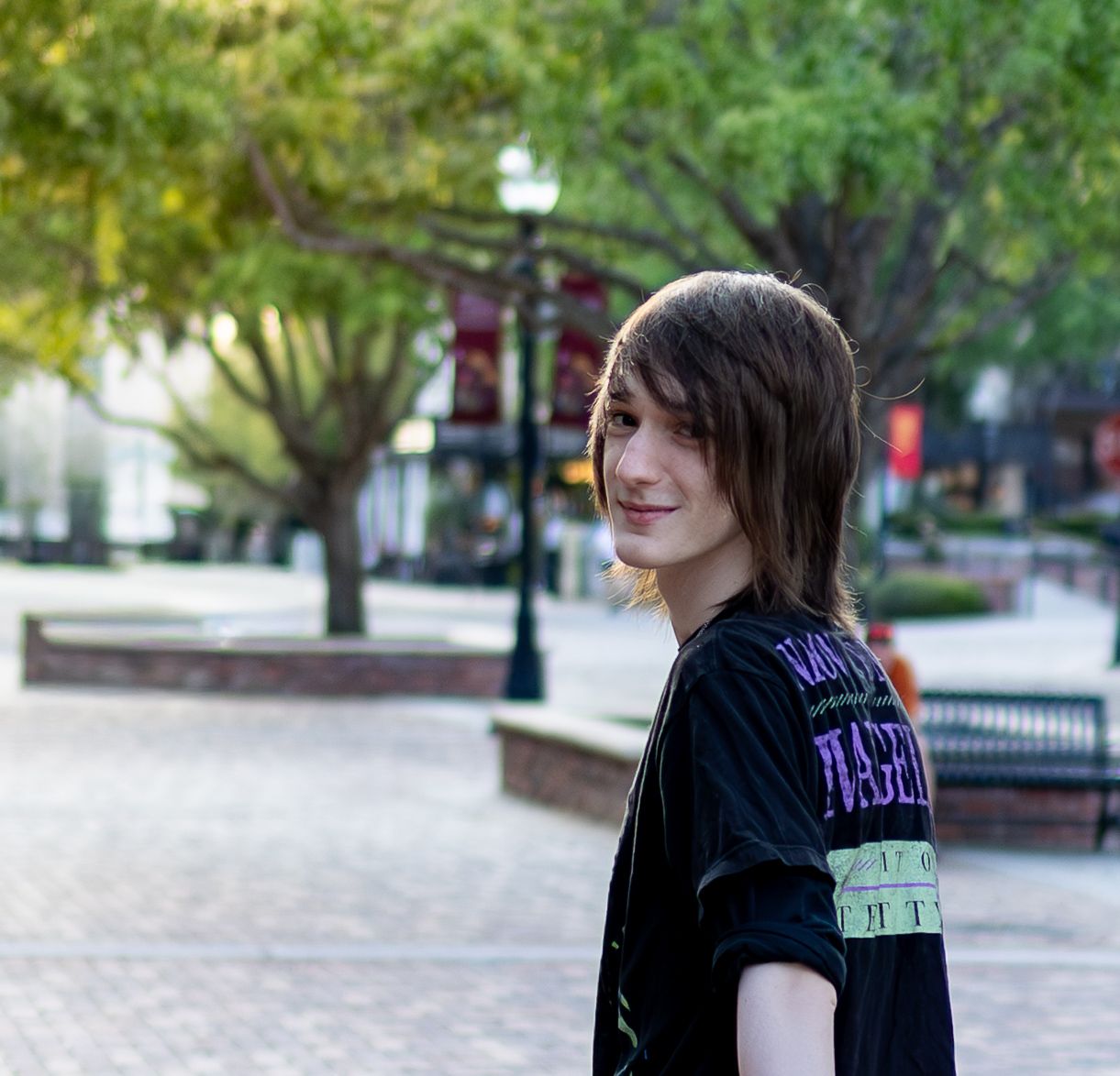A person in a black t-shirt smiling outdoors in an urban plaza with trees and lamp posts in the background.