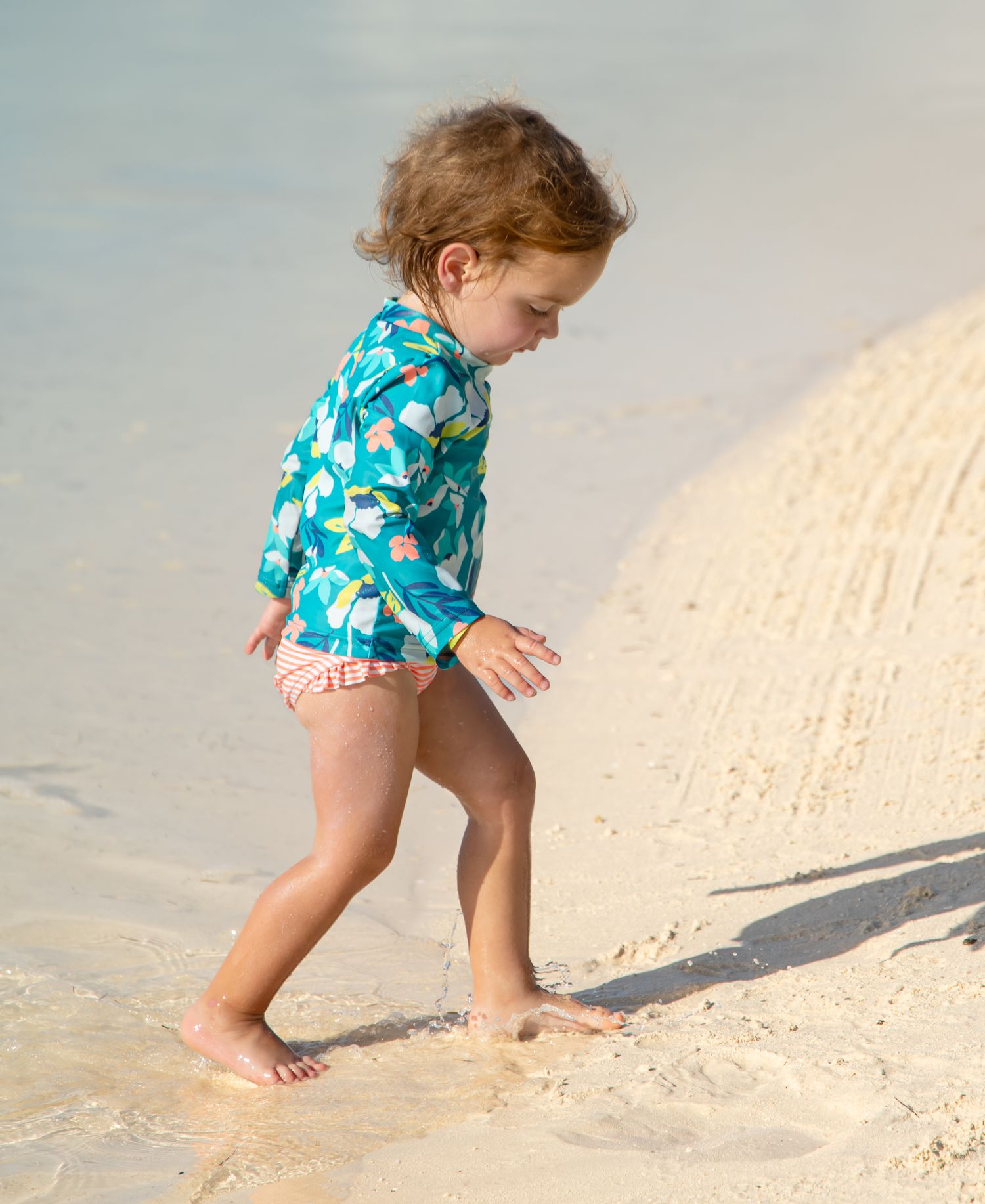 Toddler in a teal long-sleeve rash guard and pink swim bottoms walks barefoot along the wet sand at the water's edge on a sunny beach.