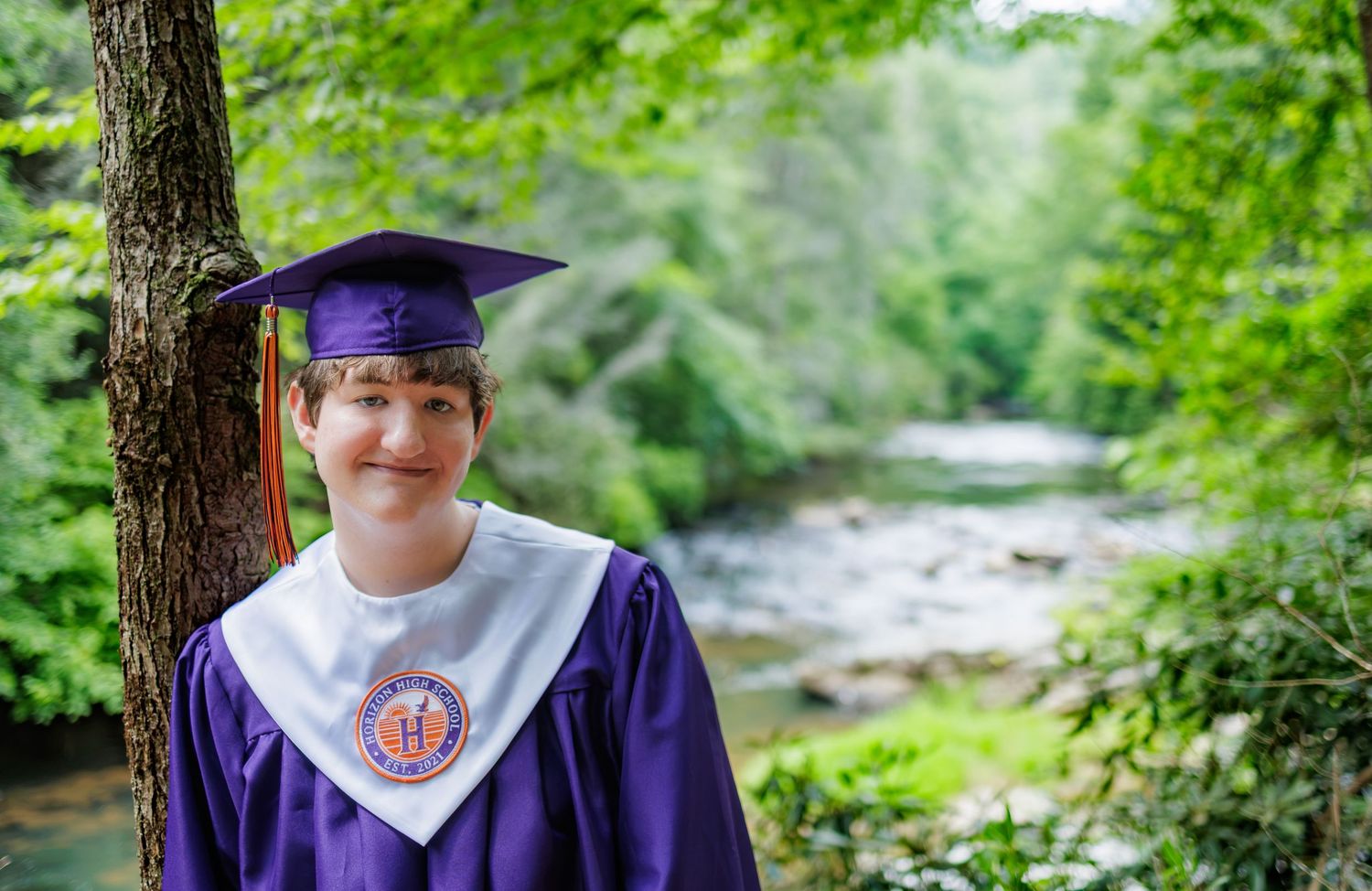 Smiling graduate in a purple cap and gown with a white stole stands beside a tree, with a forest stream and green foliage in the background.