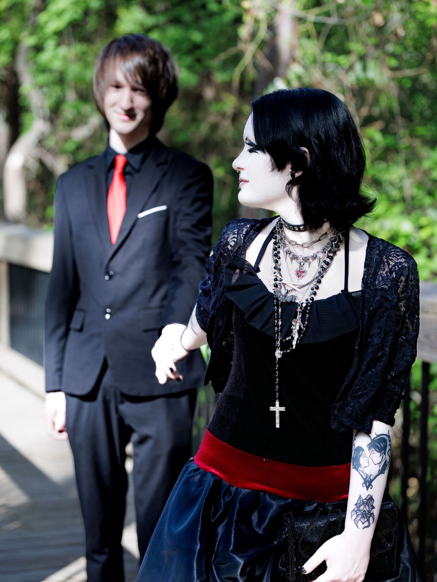 Gothic woman in black lace dress with cross necklace stands beside man in black suit with red tie on bridge.