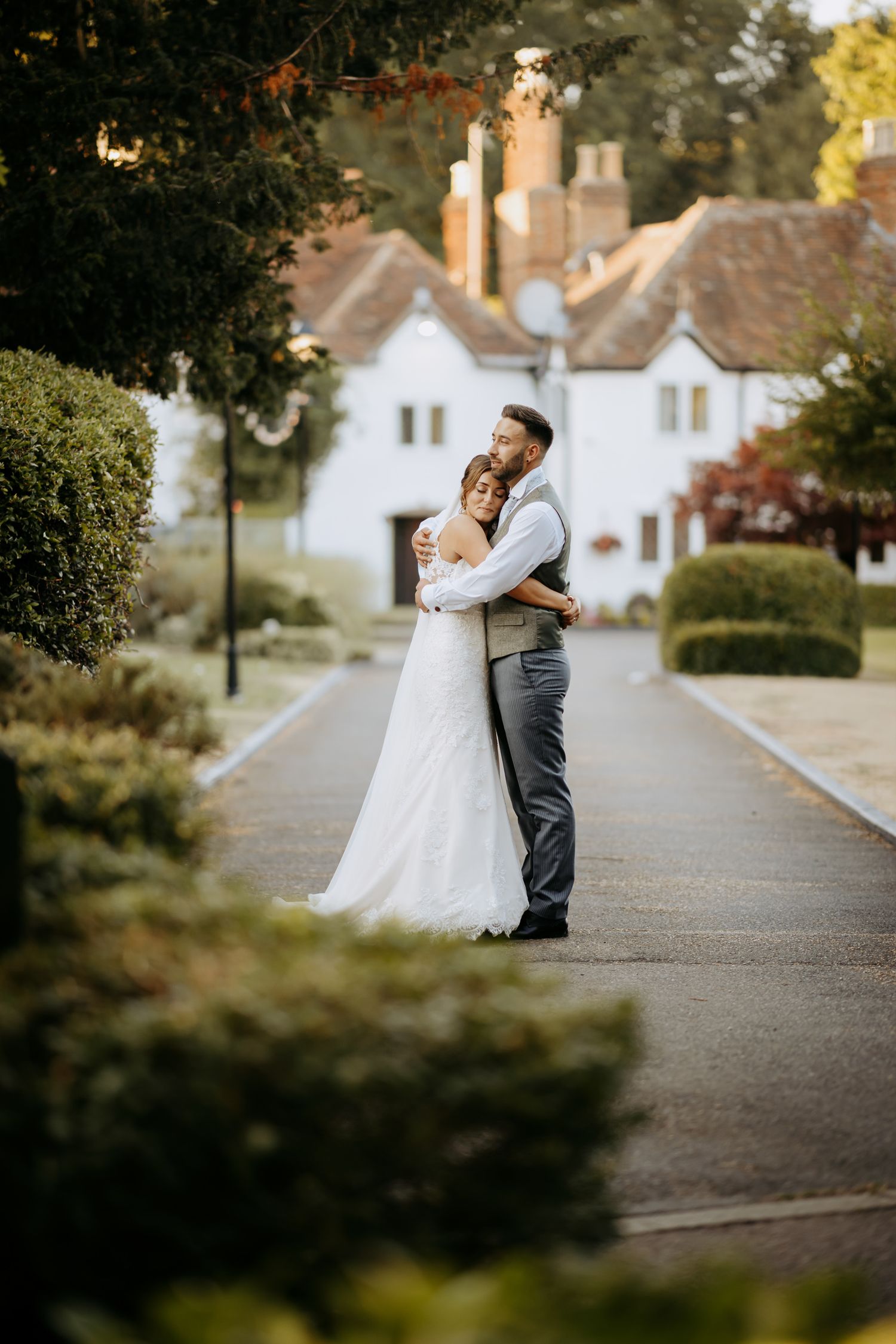 A romantic wedding couple embraces on a path with charming white cottages and autumn foliage in the background in Bedford.