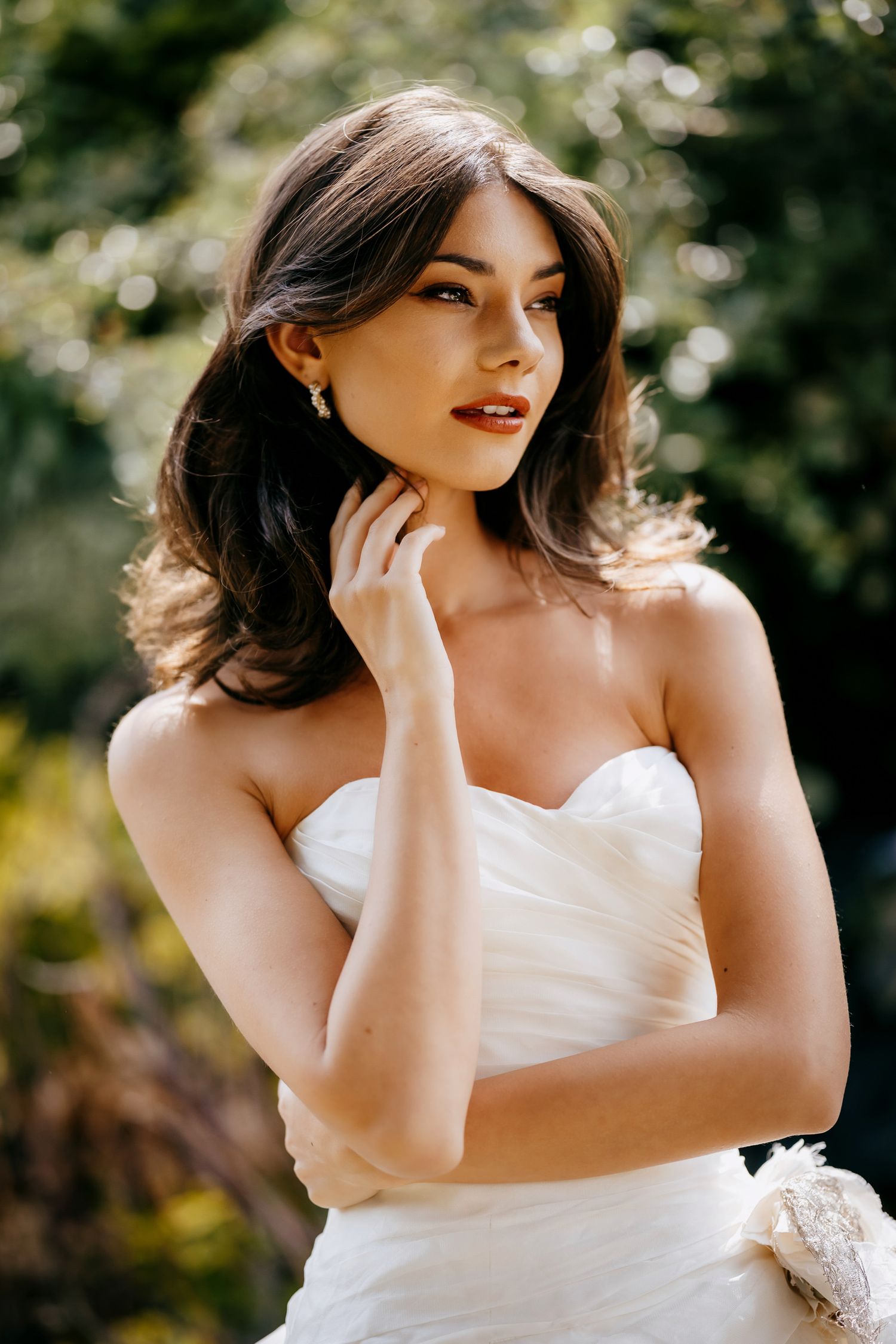 A bride in a strapless white wedding dress poses outdoors with soft natural lighting and greenery in the background.