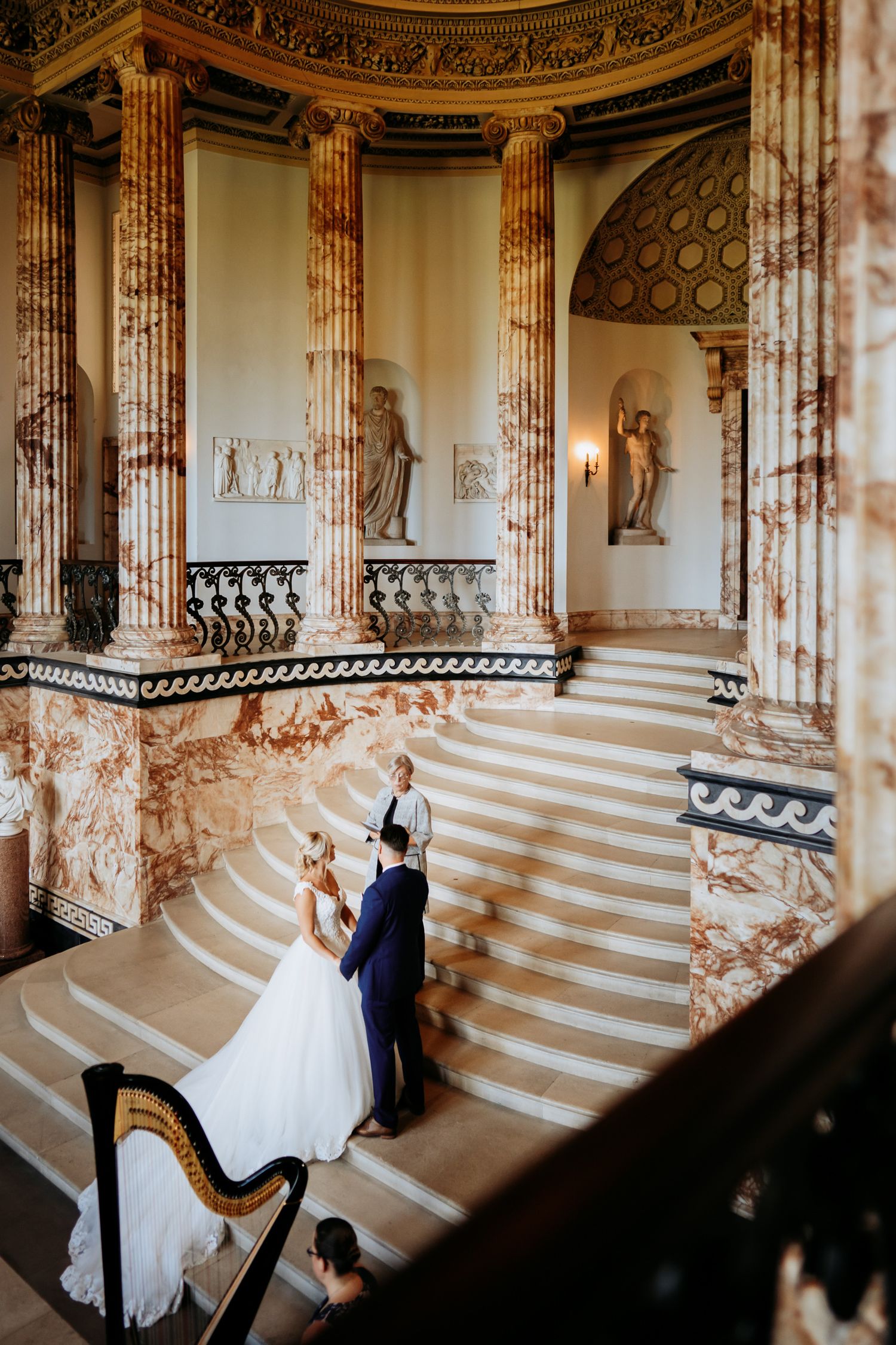 Elegant wedding couple ascends marble staircase in ornate classical interior while harp music plays during their ceremony.