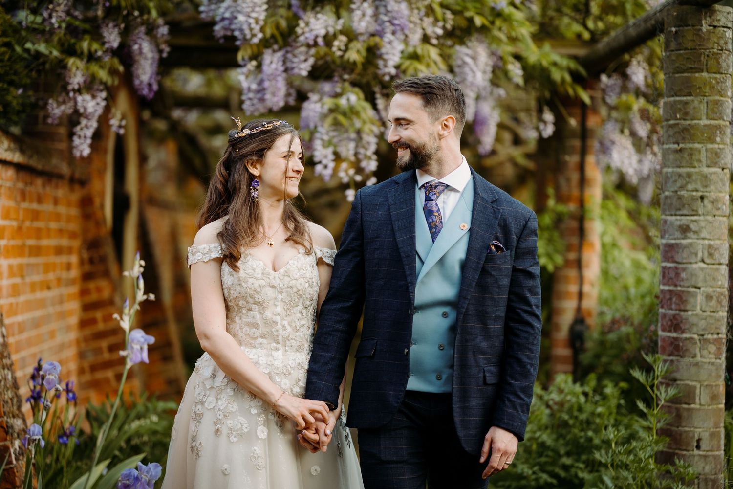 A couple shares romantic moments under blooming wisteria in a garden setting during their wedding celebration at Lanwades Hall.