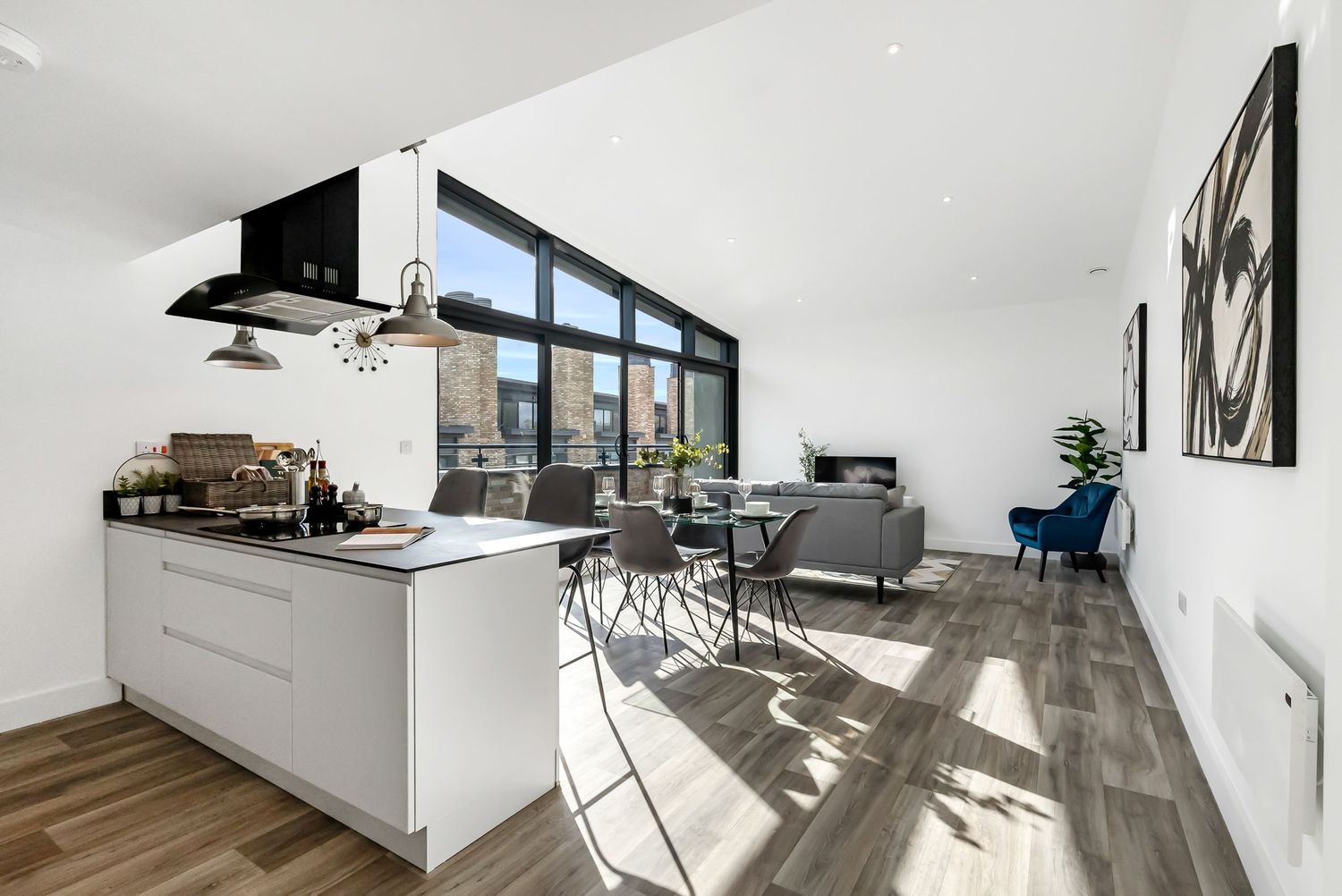 Modern apartment interior showcases sleek white kitchen, hardwood floors, and natural light streaming through large windows.