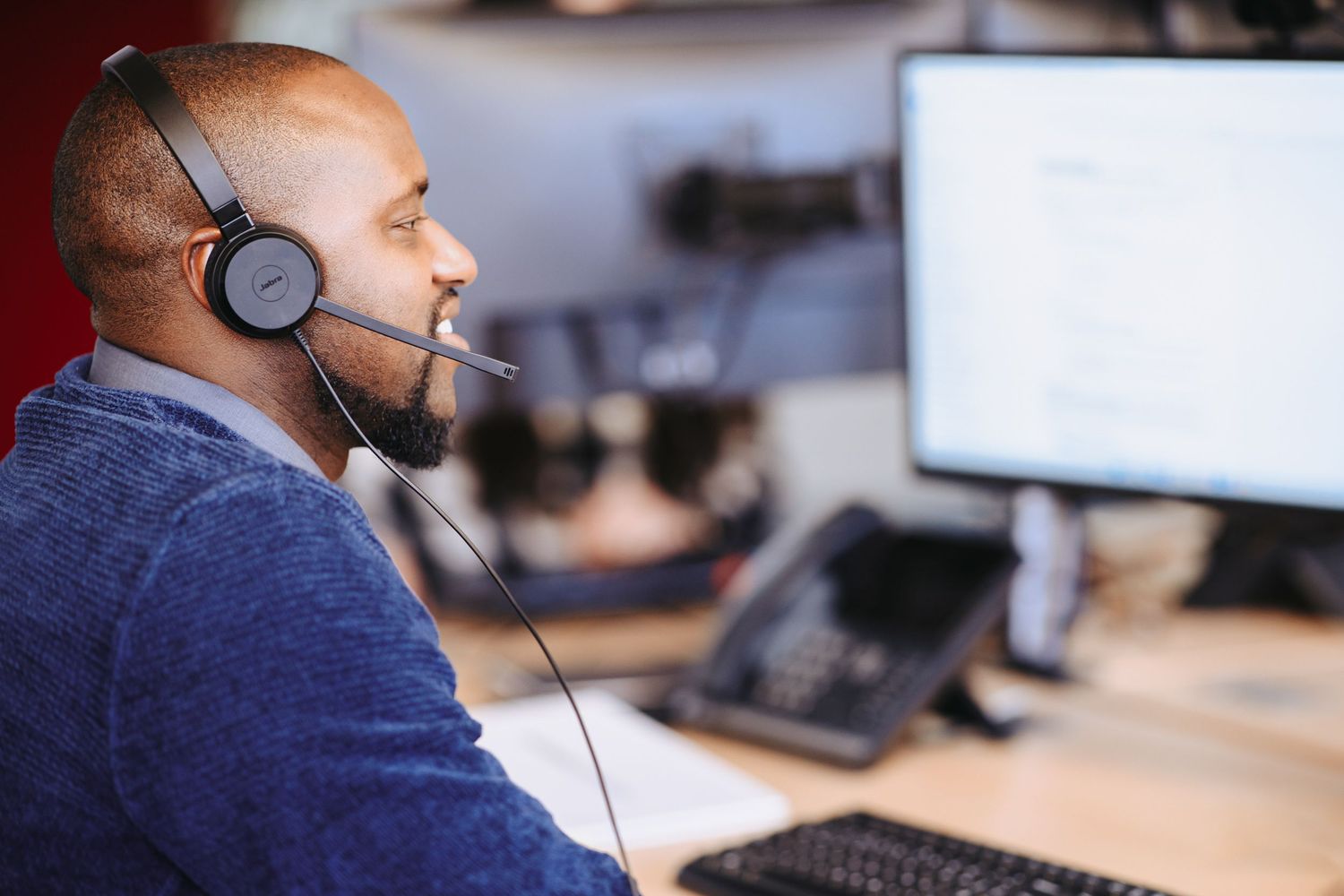Customer service representative wearing headset works at computer desk in office environment.