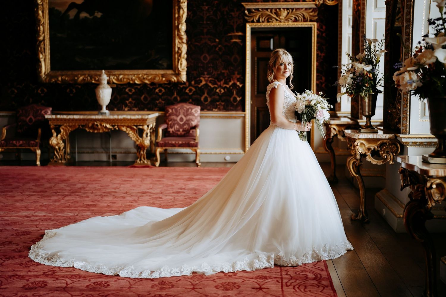 A stunning bridal portrait in an elegant room with ornate gold frames and red carpeting highlighting a beautiful long flowing wedding dress.