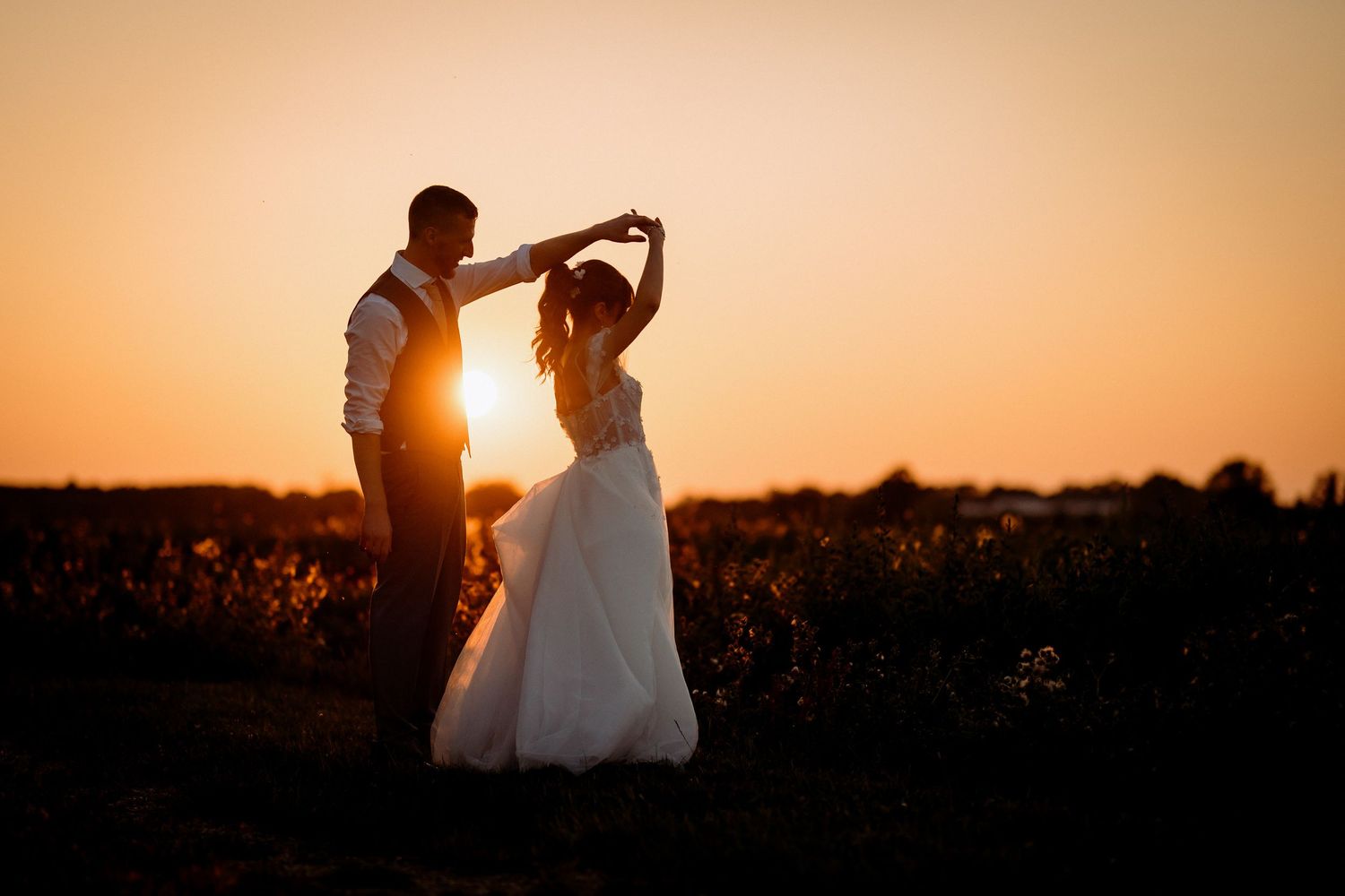 Bride and groom dancing at sunset in a golden field, silhouetted against a vibrant orange sky.