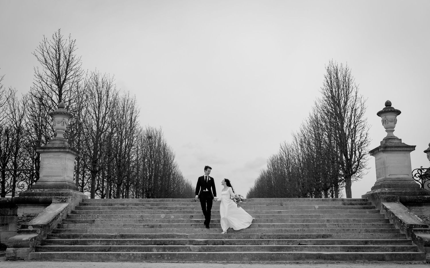 A black and white photo showing a wedding couple walking up stone steps in a park setting with bare winter trees.