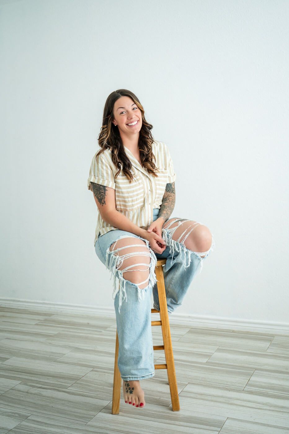 Casual portrait of woman in ripped jeans and striped shirt sitting on wooden stool.