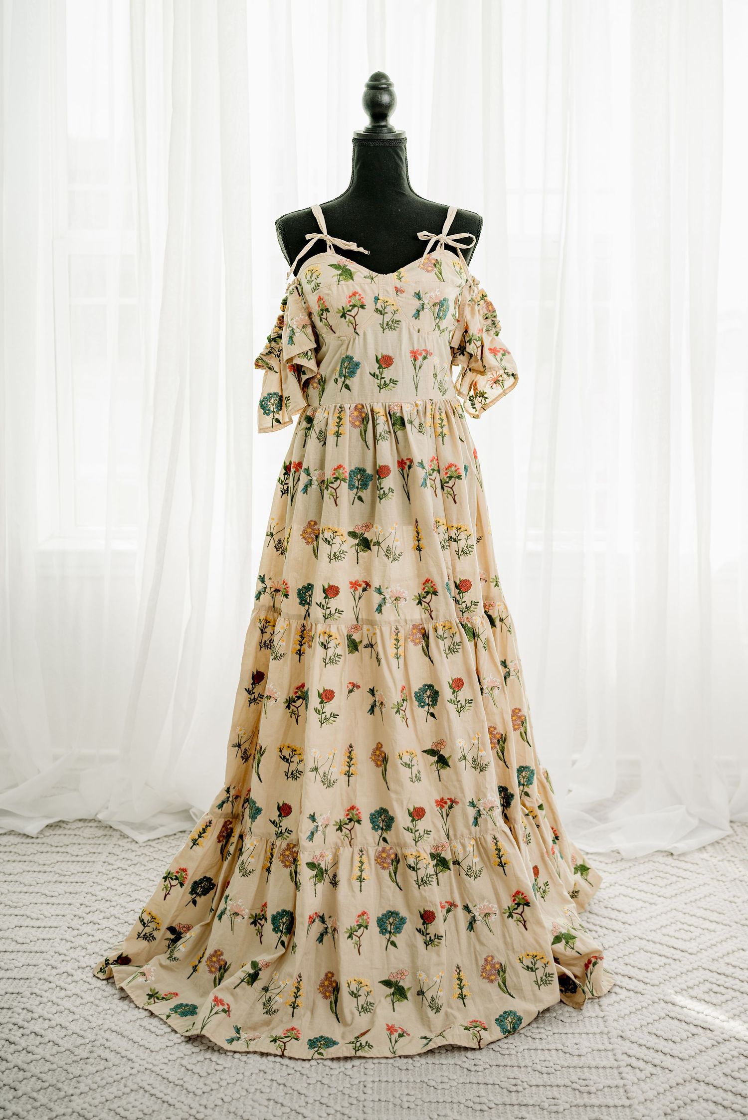 Floral print maxi dress on mannequin, cream background, tiered skirt, off-shoulder ruffle, spaghetti straps, colorful botanical pattern.