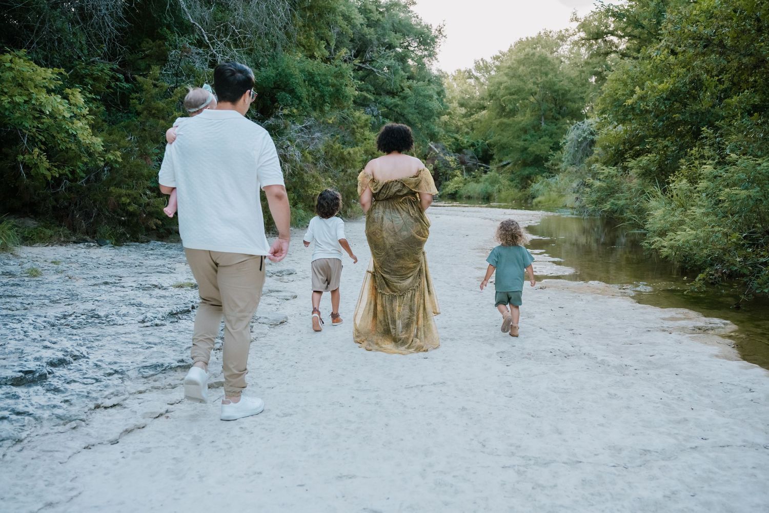 Family walking along a sandy riverbank surrounded by lush green trees in a serene outdoor setting.