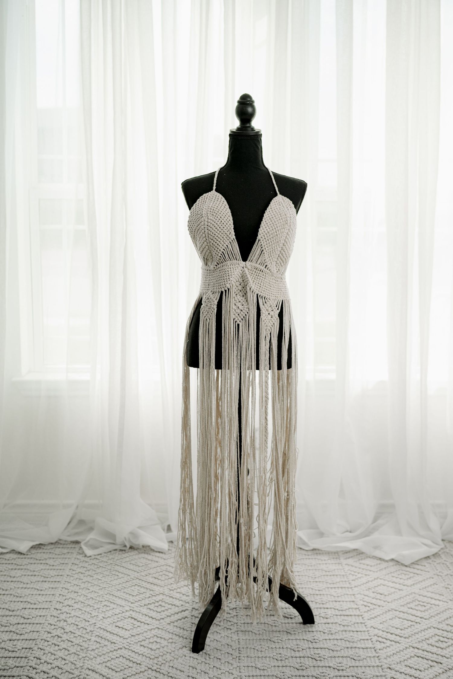 Handmade macrame halter top dress displayed on black mannequin with long fringe skirt in boho white cotton style.