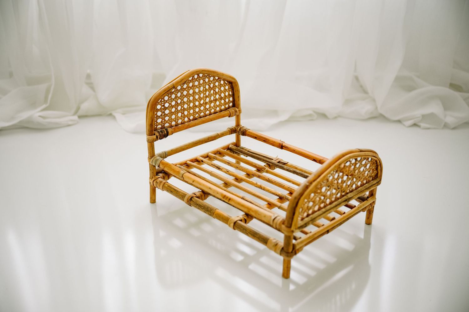 Small vintage-style rattan newborn photography prop bed with woven headboard on white reflective surface.
