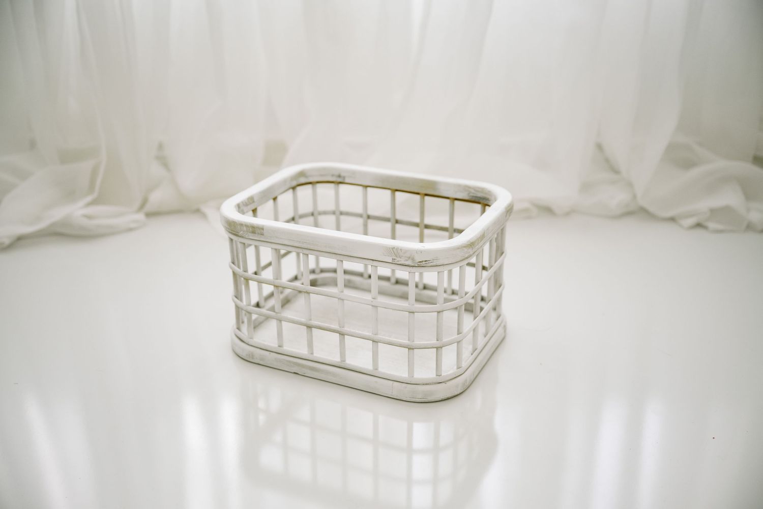 Small white plastic wicker-style storage basket on white reflective surface against sheer curtains.