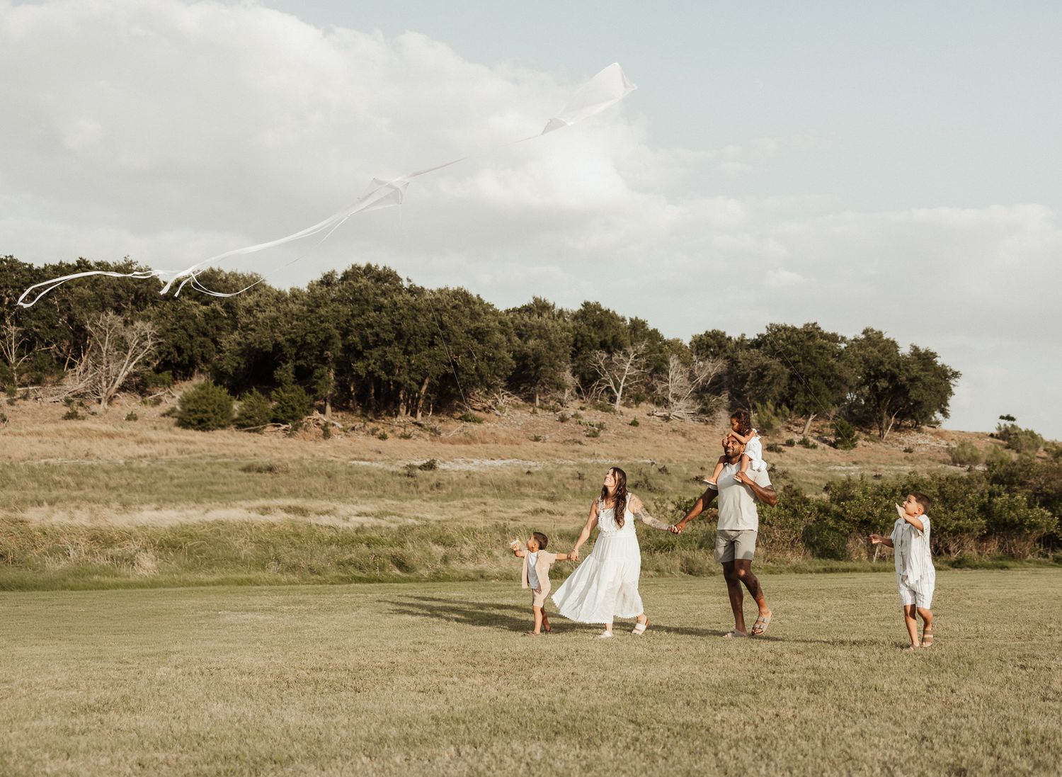 Family of four flying a kite together in an open grassy field on a cloudy day.